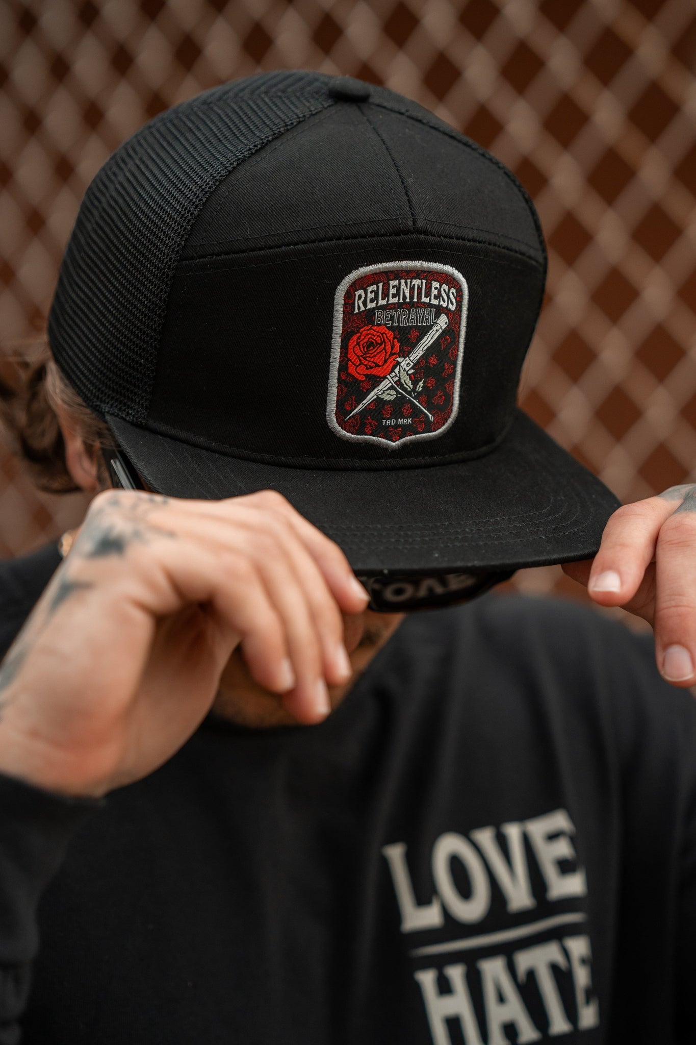 A person adjusts a black snapback from Relentless Betrayal featuring the Love/Hate 7 Panel design, with a Relentless patch showing a red rose and crossed chains. Tattooed hands adjust it, while they sport a shirt with Love and Hate. A chain-link fence is in the background.