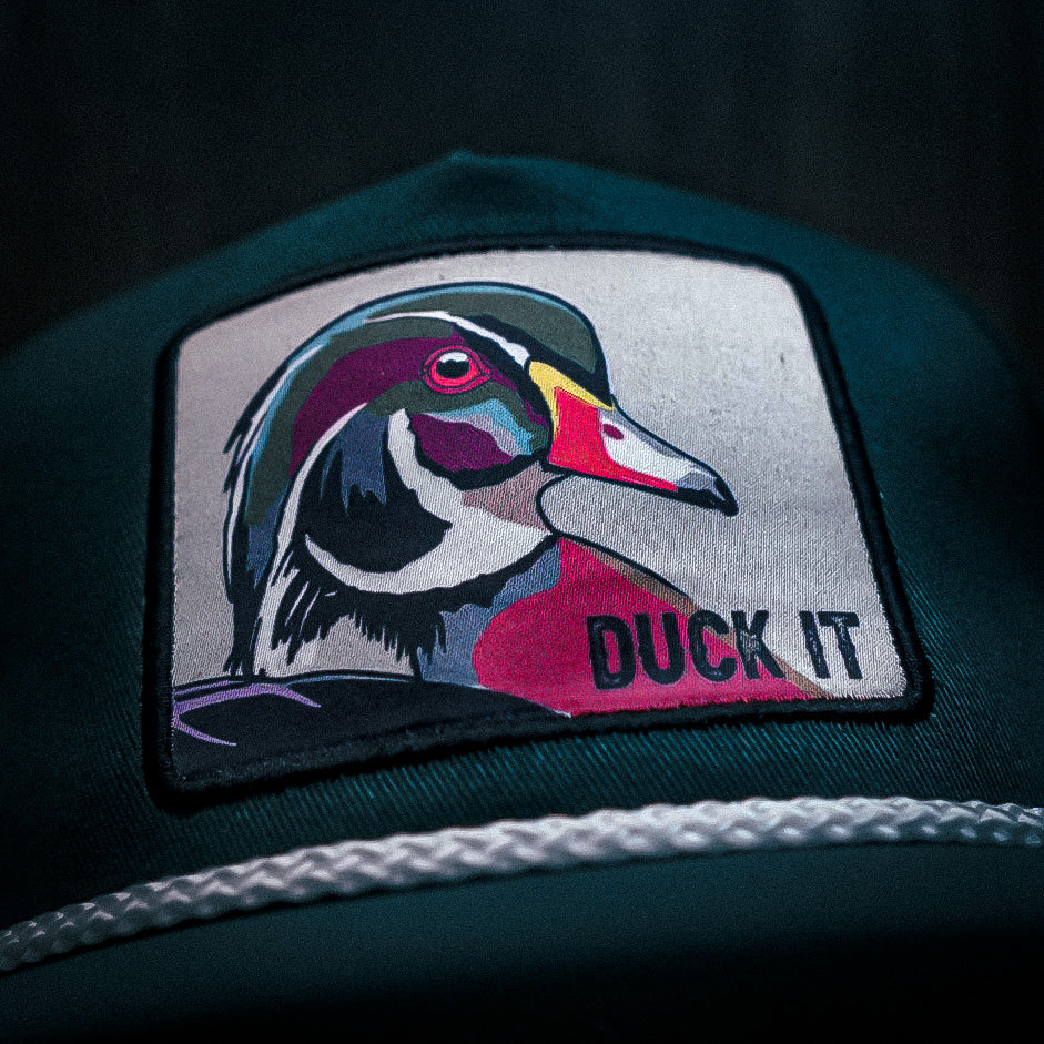 The Mucker Bucket Duck It - OG Green Wood Duck bucket hat features a handmade design with a high-quality embroidered patch of a colorful duck and DUCK IT, complemented by a white braided rope above the brim.