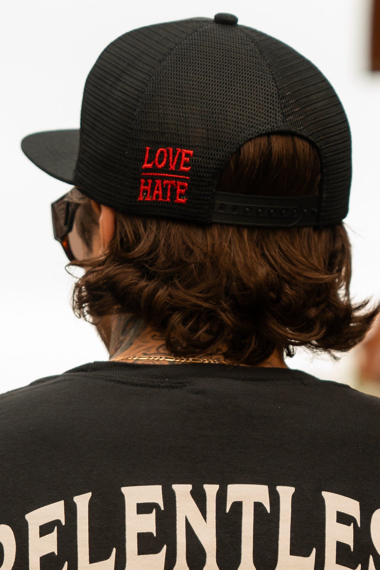 A person with shoulder-length brown hair and sunglasses wears a Love/Hate 7 Panel Trucker Snapback from Relentless Betrayal featuring red text, paired with a black shirt displaying RELENTLESS in white.