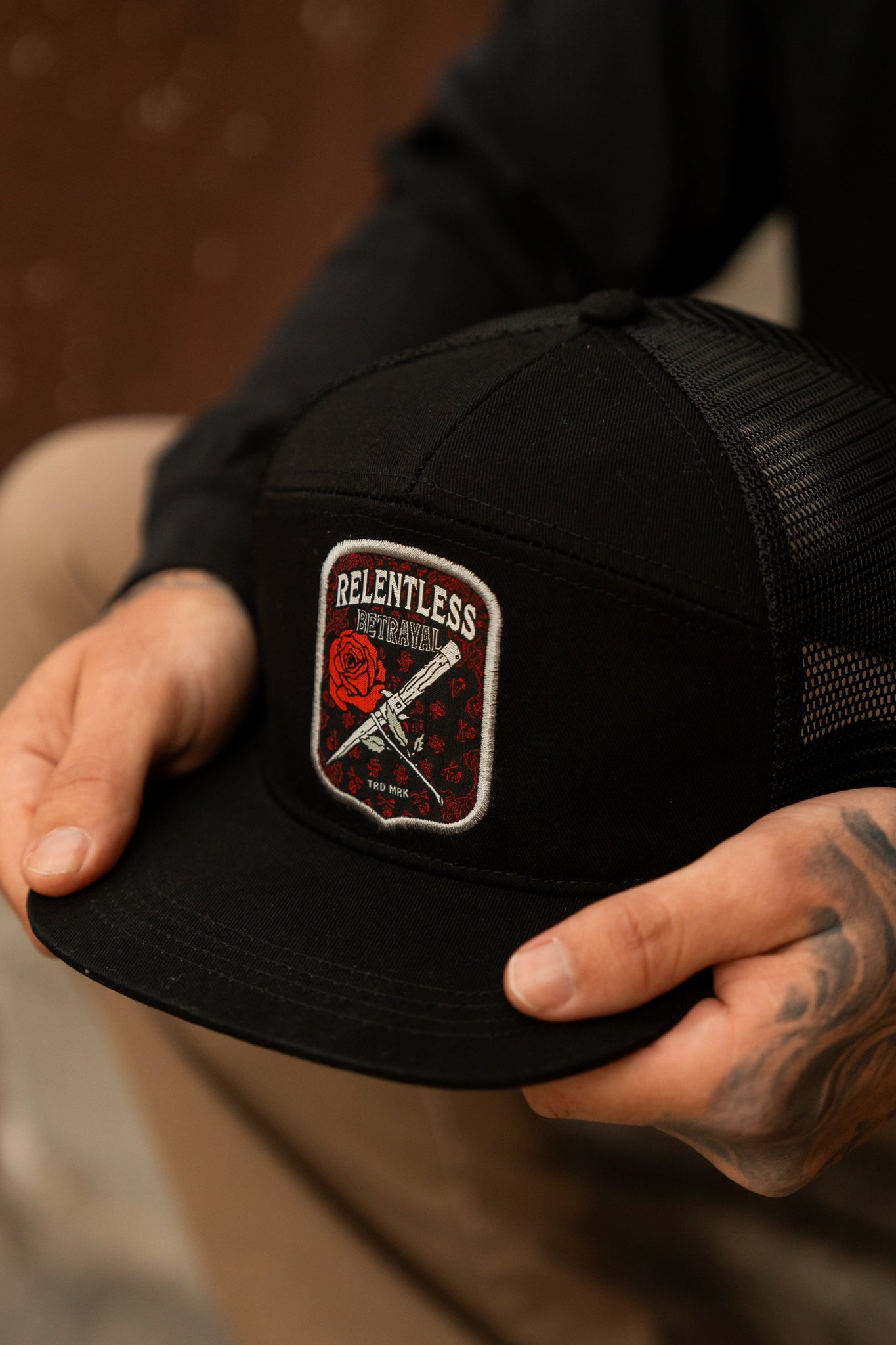 A tattooed individual holds a handmade black Love/Hate 7 Panel Trucker Snapback by Relentless Betrayal, adorned with a rose and two swords patch. Theyre wearing a black long-sleeve shirt and beige pants.
