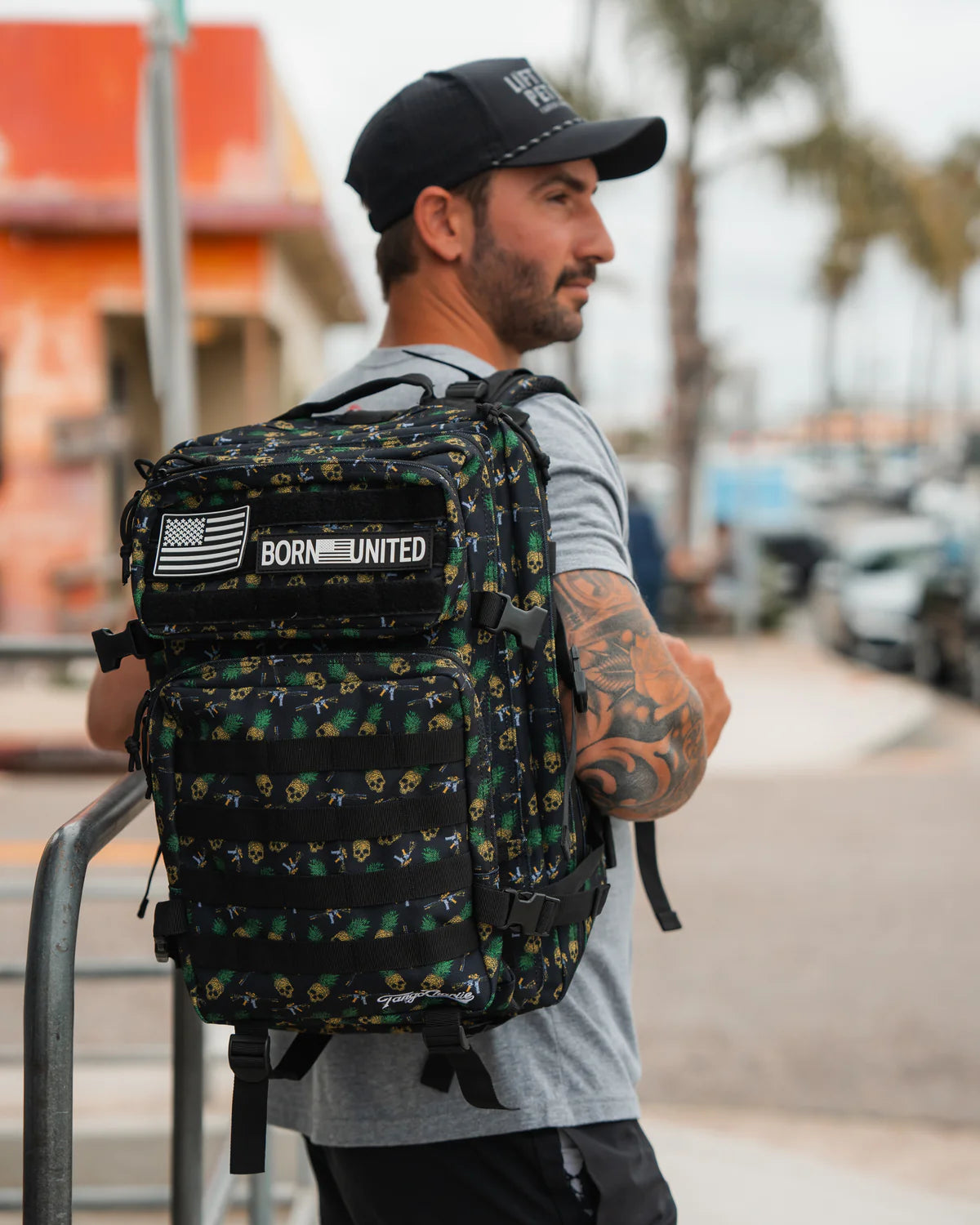 A tattooed man outdoors showcases tactical versatility with the TCA Tac-Pac - Tropic Thunder 2.0 - 45L backpack by Born United Private Label, featuring green patterns and an American flag patch. The waterproof black backpack complements his gray shirt and black cap against palm trees and blurred buildings.