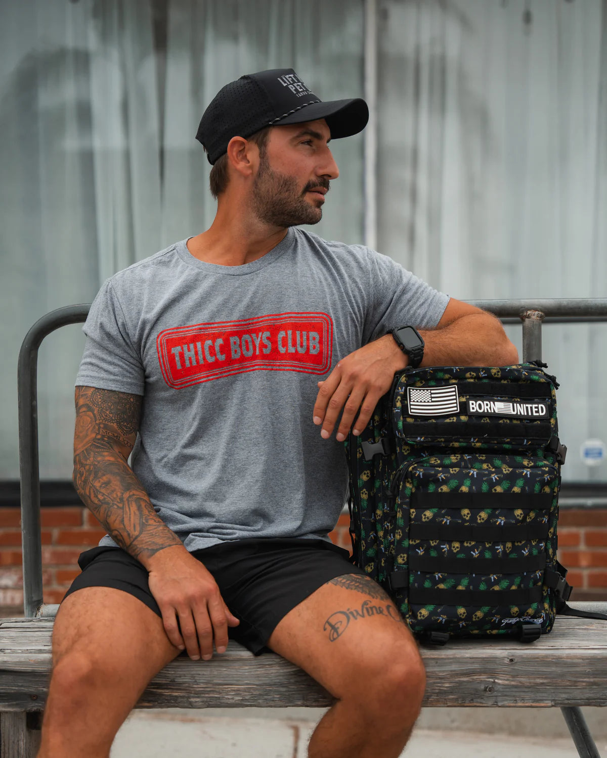 A bearded, tattooed man sits on a bench, wearing a black cap, gray Thicc Boys Club t-shirt, and black shorts. He leans on his versatile Born United Private Label TCA Tac-Pac - Tropic Thunder 2.0 - 45L (Warehouse), with patches like the U.S. flag. A large window is in the background.