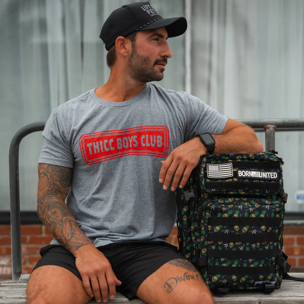A man in a black cap and gray Tango Charlie Apparel THICC Boys Club T-Shirt sits on a bench with his arm on a black backpack featuring an American flag patch, looking left—ready to lift or just showing its Time To Eat.