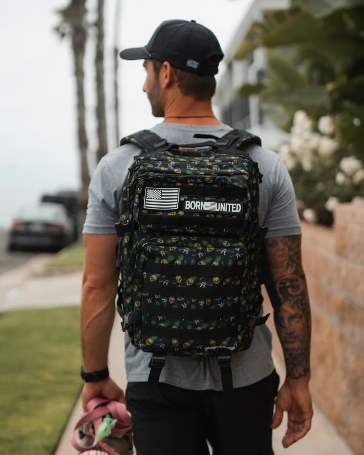 A tattooed man strolls down the sidewalk in a black cap and gray t-shirt, carrying a TCA Tac-Pac - Tropic Thunder 2.0 (45L) from Born United Private Label, complete with an American flag and Born United patch. Pink and green items are in his left hand against trees and a wall backdrop.