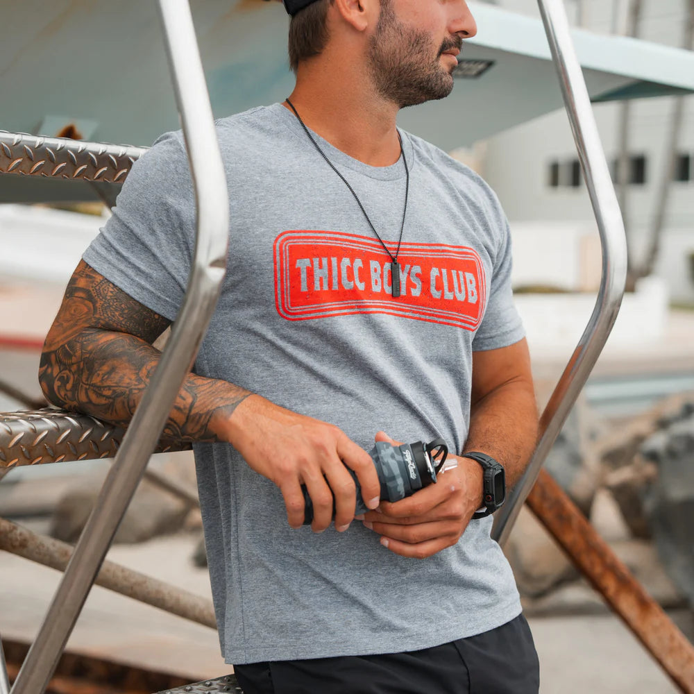 A bearded, tattooed man in a gray Tango Charlie Apparel THICC Boys Club T-Shirt and black cap leans on metal stairs with a camera lens, standing outdoors by boats—ready to lift after getting the shot.