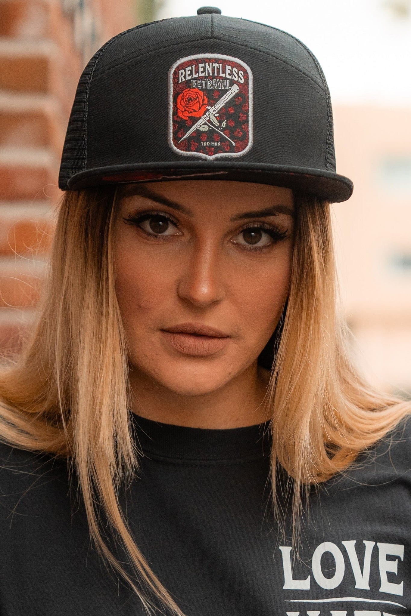 A person with long blonde hair wears a Relentless Betrayal Love/Hate 7 Panel Trucker Snapback and a partially visible black shirt. The backdrop is a blurred mix of brick wall and greenery.