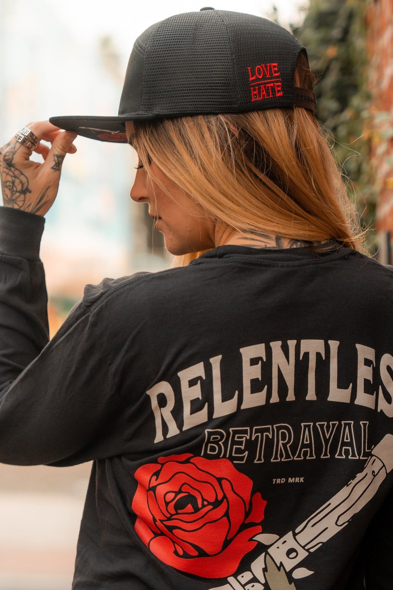 A person turned away, wearing a handmade Love/Hate 7 Panel Trucker Snapback with LOVE HATE in red and a Relentless Betrayal black shirt featuring RELENTLESS BETRAYAL above a vivid red rose and knife design, all enhanced by tattoos on their hand and neck for an edgy look.