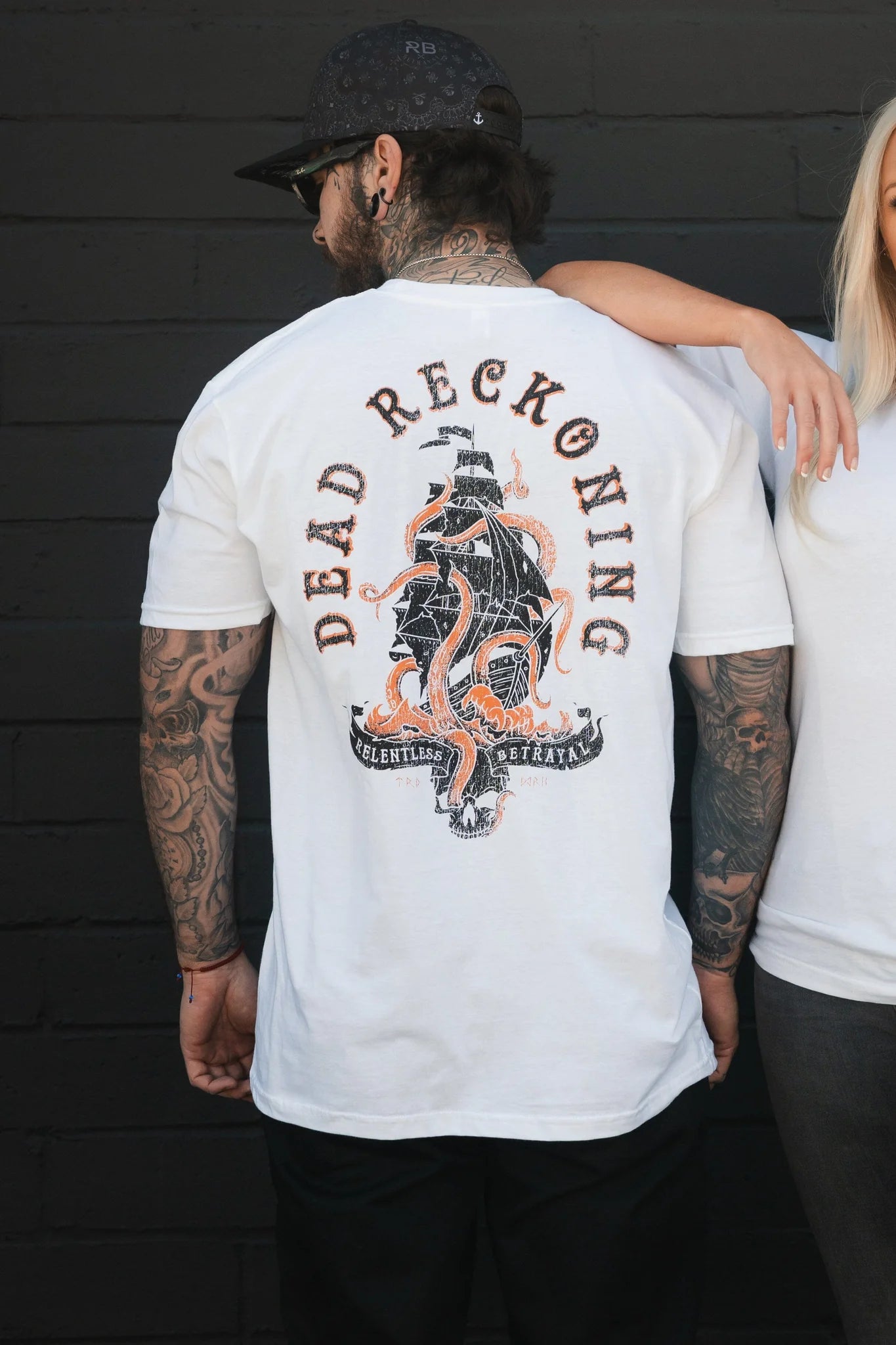 Against a dark wall, a tattooed man in the Dead Reckoning Tee- White by Relentless Betrayal is embraced by a woman. The shirt displays a detailed ship surrounded by flames and the words DEAD RECKONING, symbolizing an ancient seafaring navigation technique.