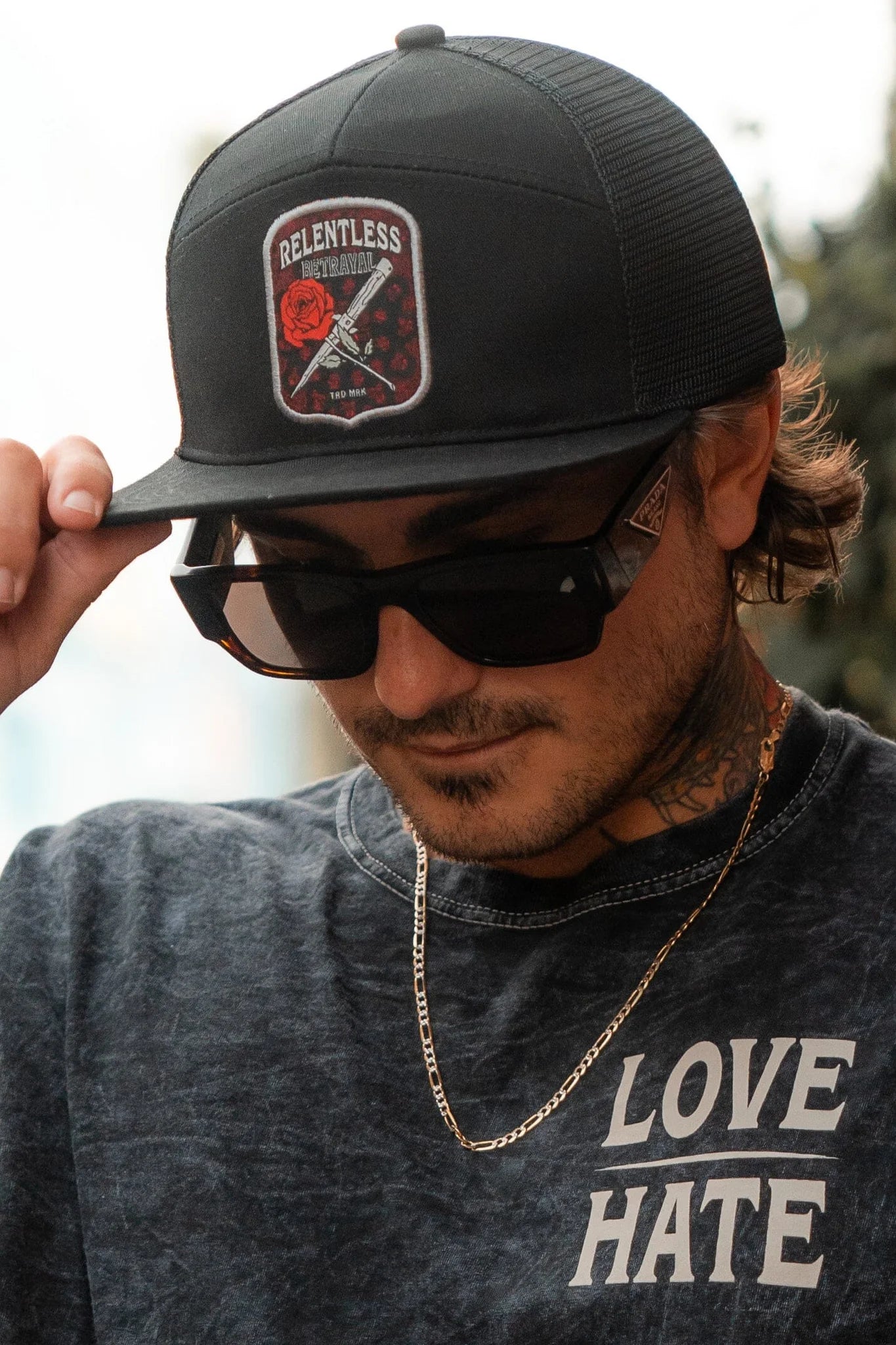 A person sports a Relentless Betrayal Love/Hate 7 Panel Trucker Snapback, sunglasses, and a dark shirt with LOVE HATE. They adjust their cap, wearing a chain necklace while tattoos embellish their neck and wrist for an edgy look.