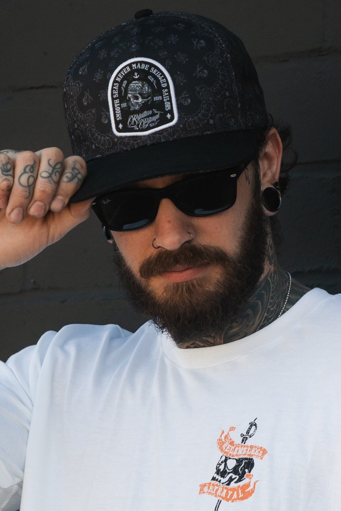A tattooed, bearded man sports sunglasses and a white T-shirt with a design. Wearing the Skilled Sailor 5th Anniversary Snapback by Relentless Betrayal, he confidently adjusts his cap brim while facing forward, embodying the resilience to overcome challenges.