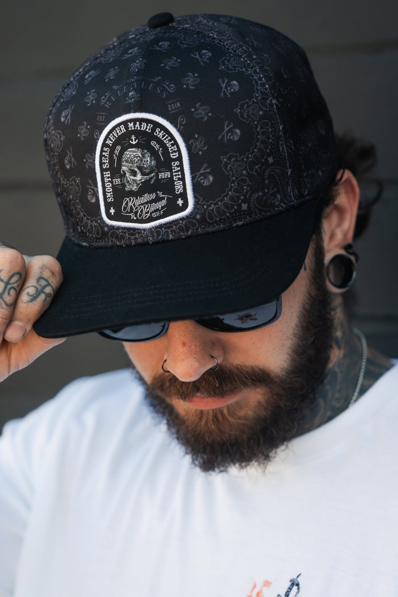A resilient man with a beard wears the Skilled Sailor 5th Anniversary Snapback by Relentless Betrayal, featuring intricate patterns and an inspiring slogan. He pairs it with sunglasses and a white shirt; tattoos cover his hand, while piercings highlight his ear and nose.