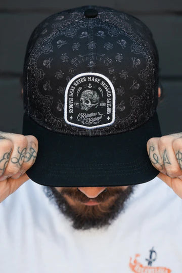 Wearing the Skilled Sailor 5th Anniversary Snapback by Relentless Betrayal, featuring intricate patterns and a front patch with text and design, this tattooed person in a white logo shirt embodies resilience and triumph over challenges.