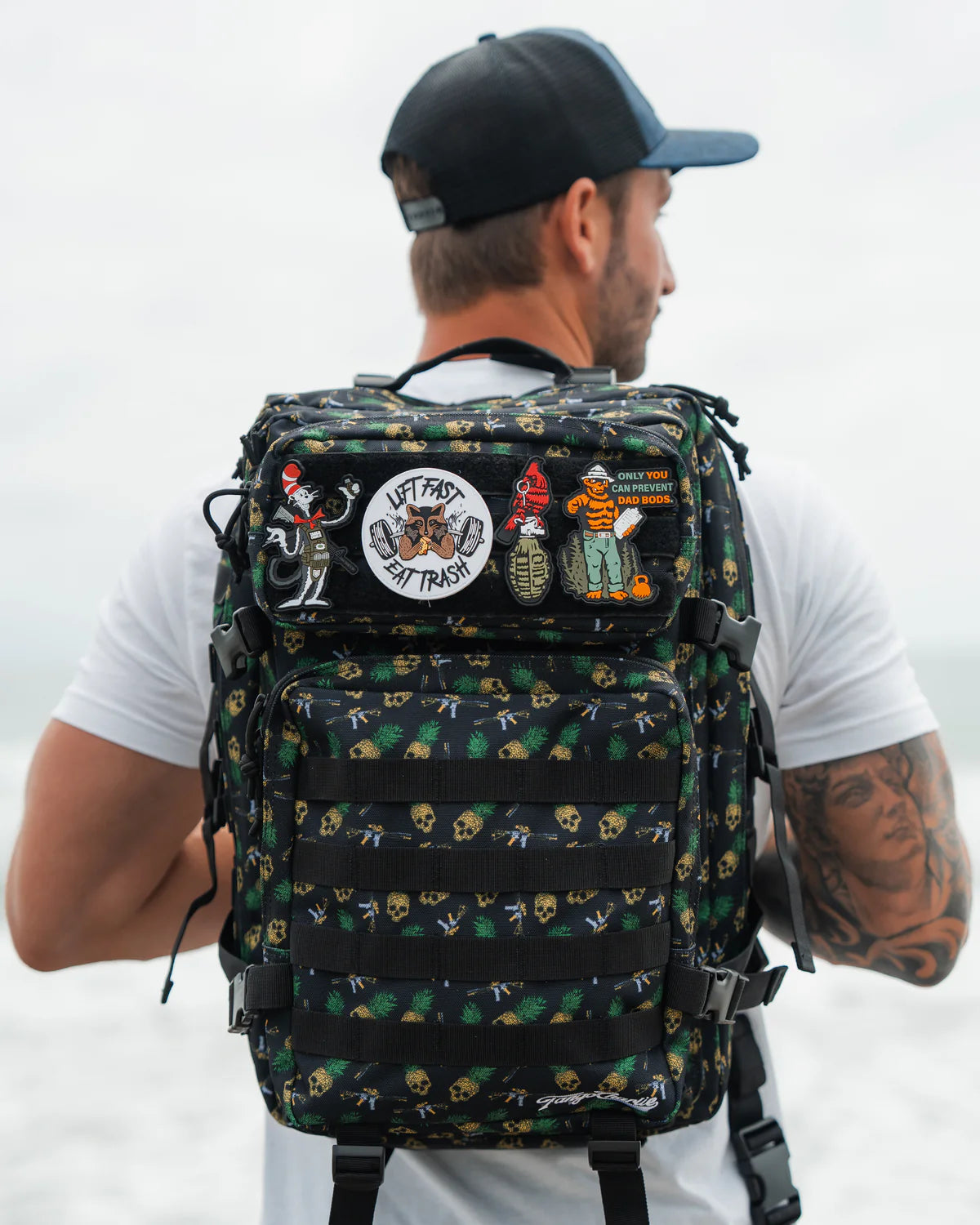 A man in a black cap and white t-shirt carries the TCA Tac-Pac Tropic Thunder 2.0 backpack by Born United Private Label, featuring colorful skull and beach-themed patches for tactical versatility, against a blurred sand and ocean backdrop.