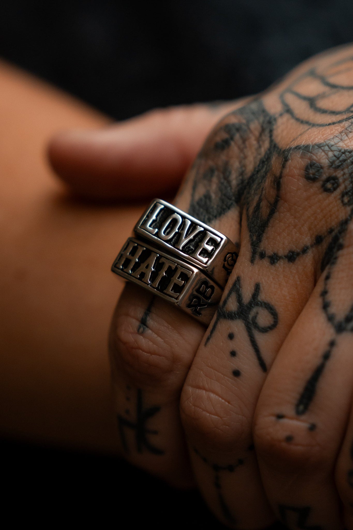 A tattooed hand, elegantly poised and adorned with Relentless Betrayals Love/Hate Rings—one spelling LOVE, the other HATE—stands out amid intricately tattooed fingers against a blurred background.