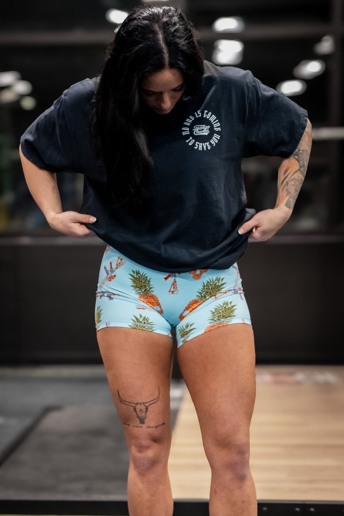 In a dimly lit gym, a woman wears Tango Charlie Apparels Womens High-Waisted Seamless Front Compression Training Shorts in Tropic Thunder. The breathable fabrics pineapple and palm tree pattern complements her bull skull tattoo on her left thigh.