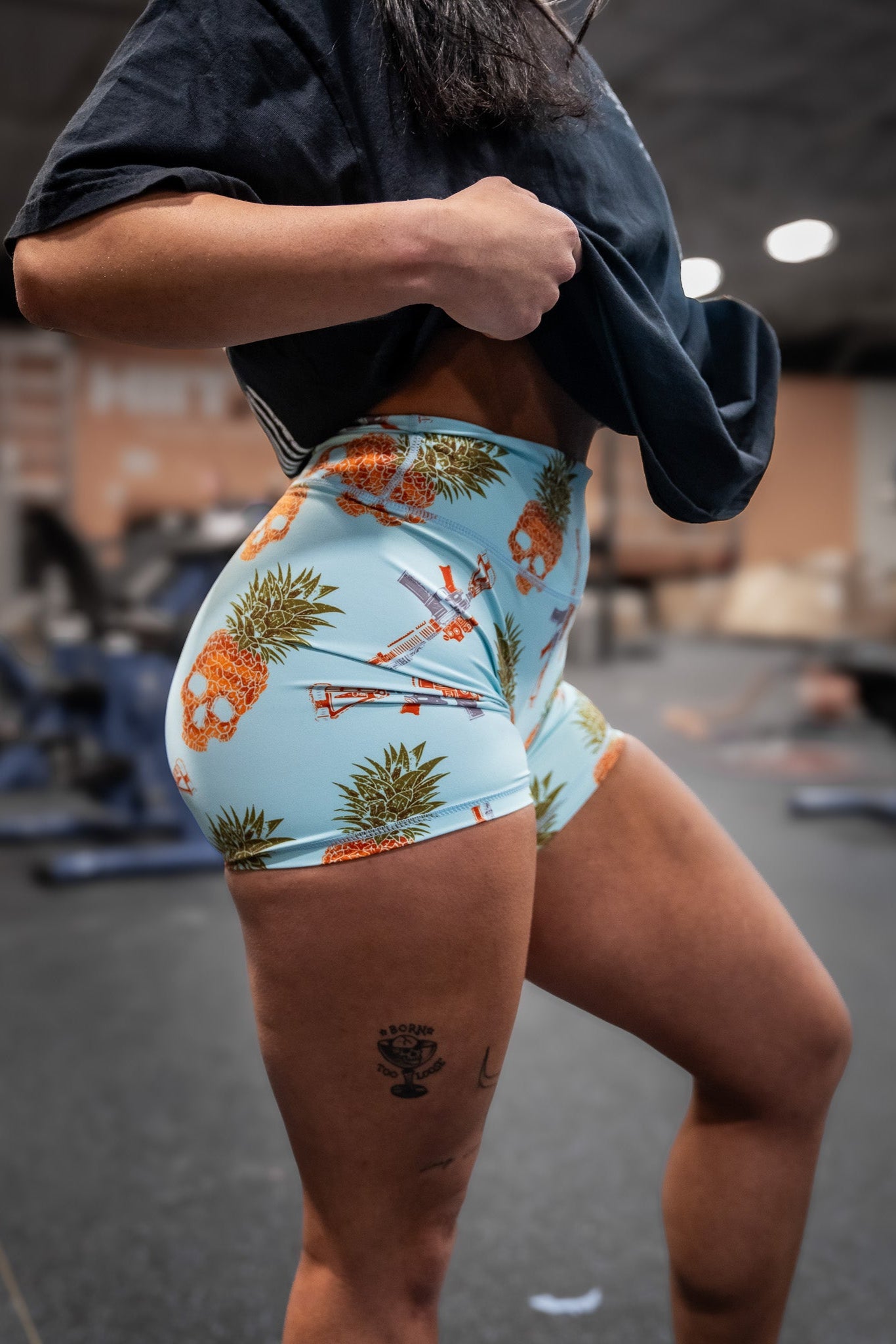 In a gym, someone lifts their black shirt, revealing Tango Charlie Apparels blue Womens High-Waisted Seamless Front Compression Training Shorts in the Tropic Thunder design with a pineapple and skull pattern. Their tattooed left thigh is set against blurred gym equipment.