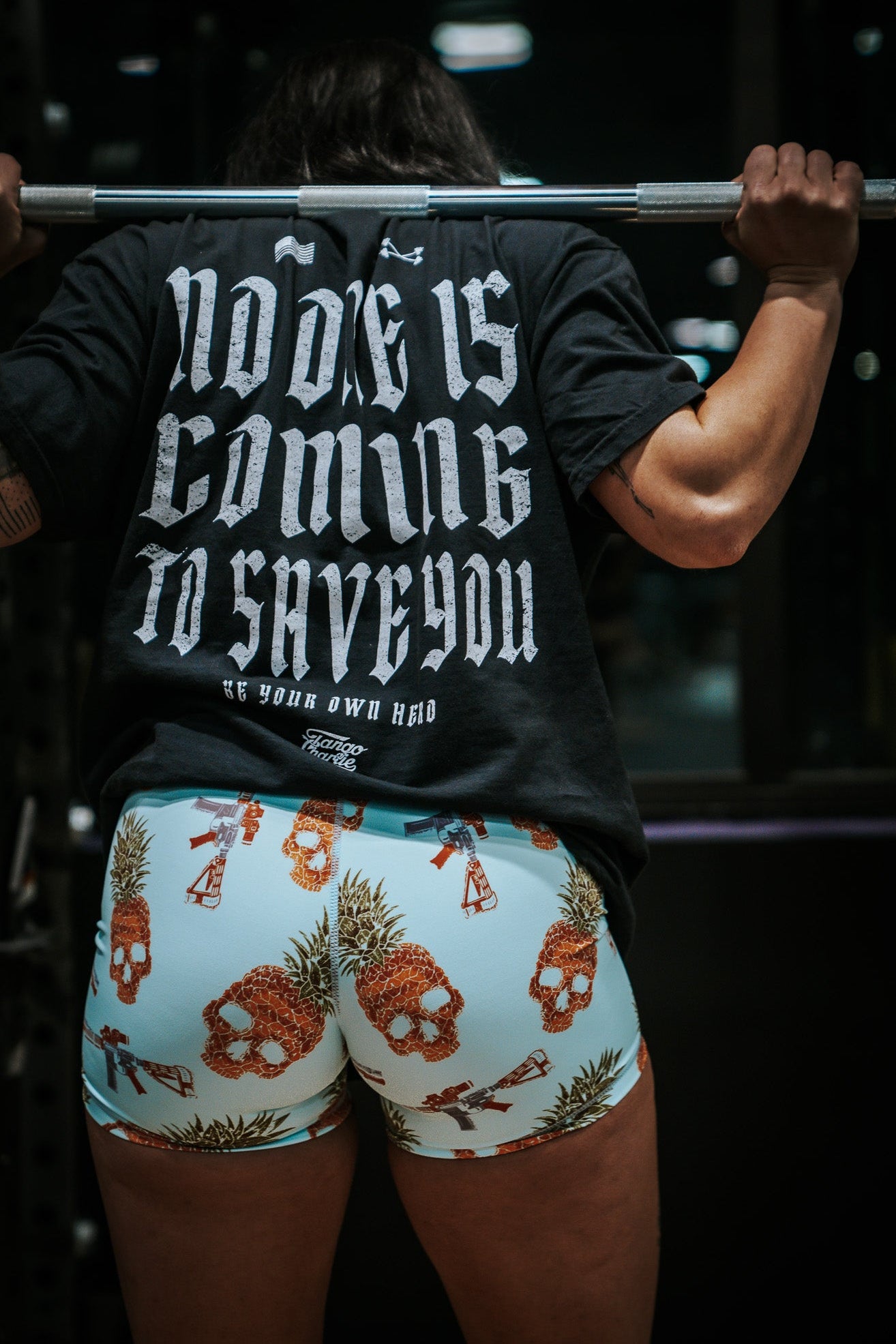 A person lifts a barbell, wearing a black shirt with bold white text: Nobody is coming to save you. Be your own hero. Their Tango Charlie Apparel Tropic Thunder shorts, featuring breathable fabric for comfort, add to the intensity of the dimly lit scene.