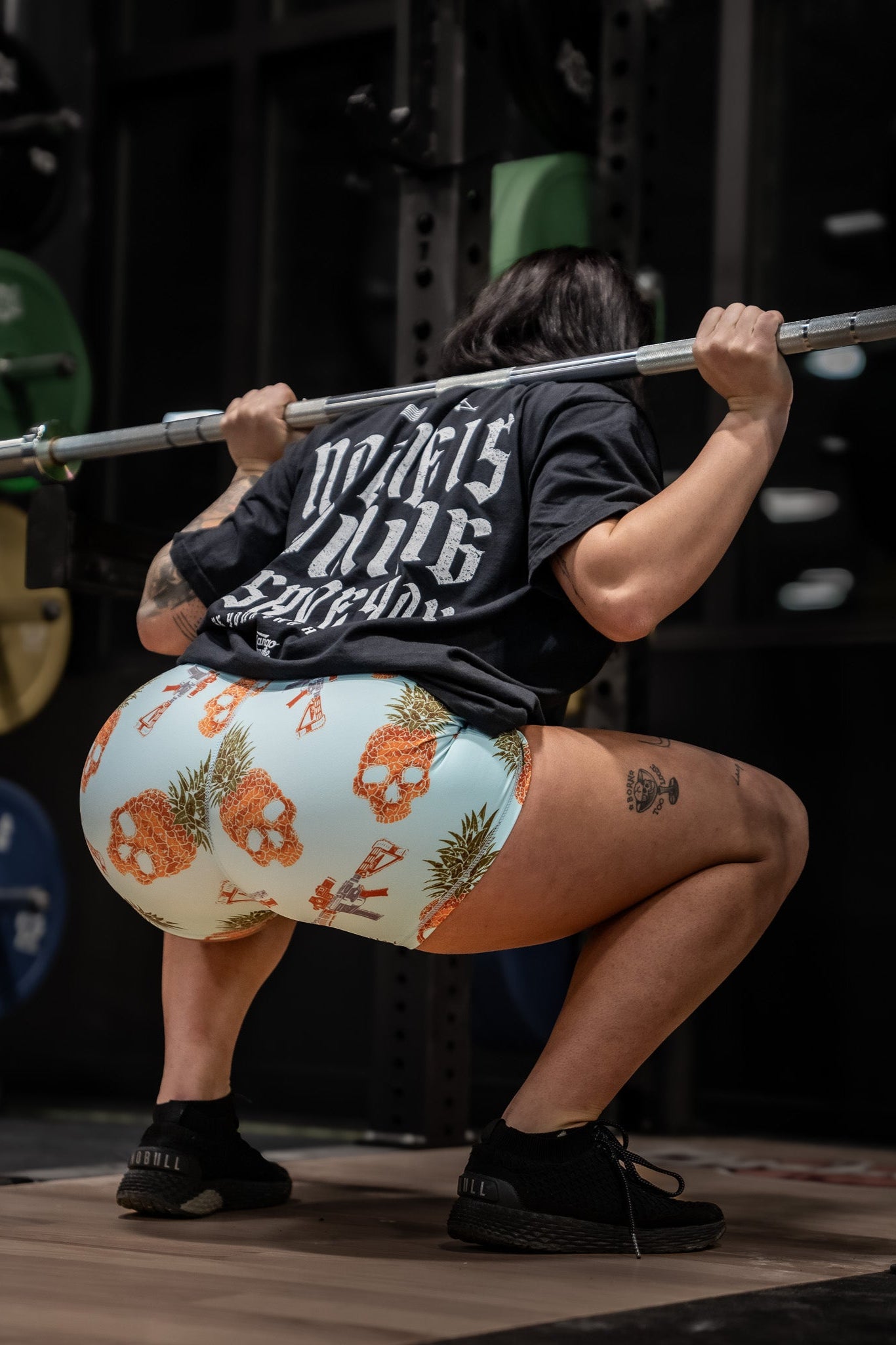 In the gym, a person does squats with a barbell on their shoulders, wearing a black shirt and Tango Charlie Apparels Womens High-Waisted Seamless Front Compression Training Shorts in Tropic Thunder. Surrounded by equipment, their gear ensures optimal comfort through each rep.