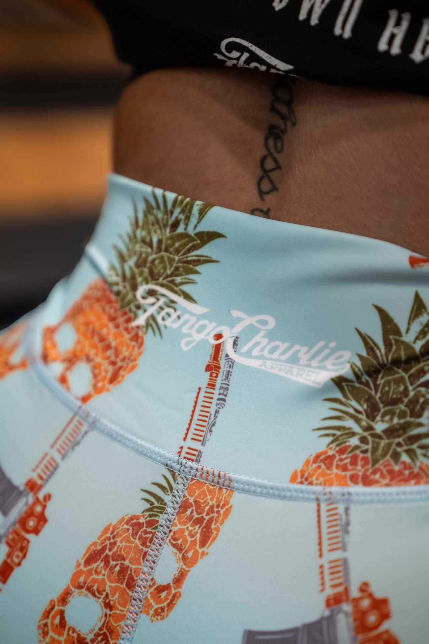 A close-up shows a persons lower back in high-waisted seamless blue shorts with orange pineapple patterns and gun illustrations. The waistband features Tango Charlie Apparel branding on these breathable fabric shorts, revealing a hint of a tattoo above.