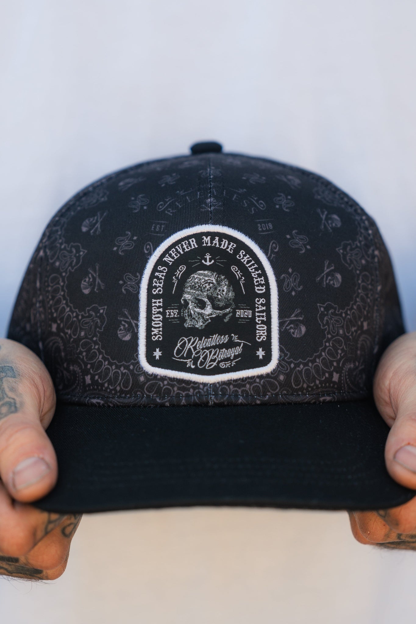 A resilient individual holds a chic black Skilled Sailor 5th Anniversary Snapback hat with decorative patterns and a patch stating Smoker 338 Never Made Sacred Blades and Relentless by Design. Tattoos on their fingers hint at the challenges theyve faced, echoing the style of Relentless Betrayal.