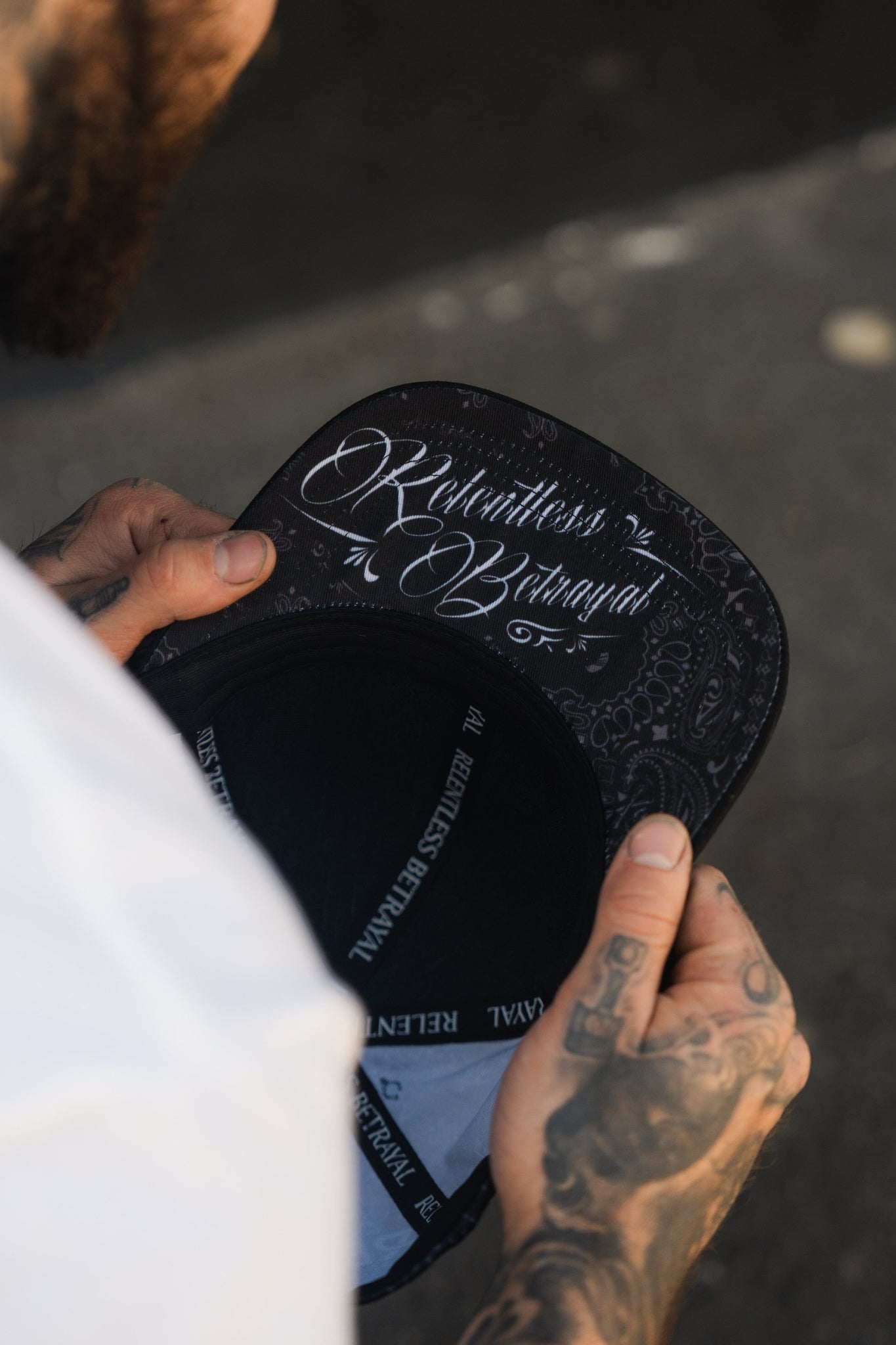 A person with tattooed hands holds the Skilled Sailor 5th Anniversary Snapback by Relentless Betrayal. The black hats underside features intricate white lettering and paisley patterns reading Relentless in Bathing, symbolizing resilience against challenges and obstacles.