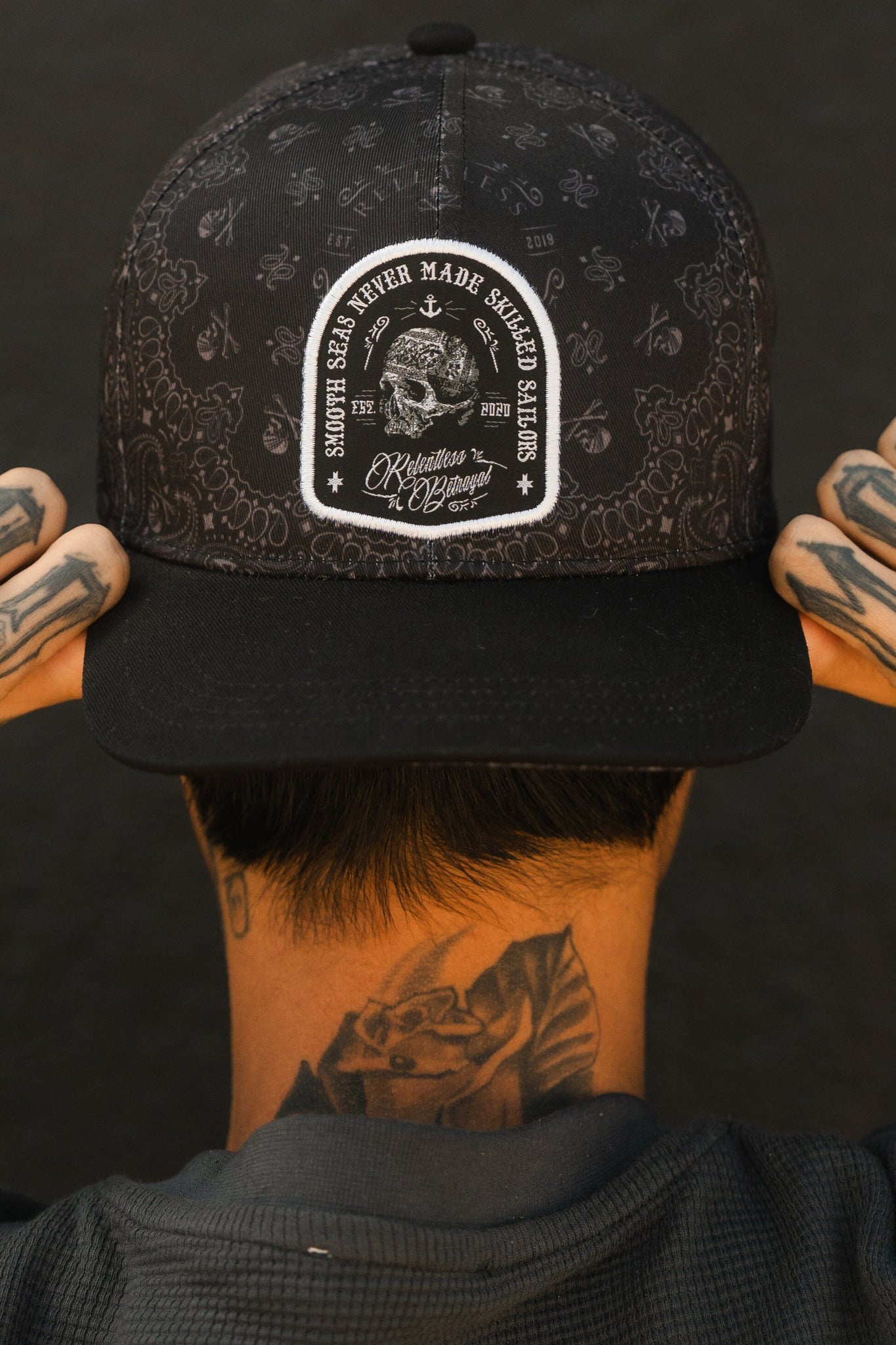 A resilient person with tattoos confidently adjusts a Skilled Sailor 5th Anniversary Snapback by Relentless Betrayal. The black hat features intricate designs with a patch reading Smooth Seas Never Made Skilled Sailors and a ship illustration symbolizing overcoming challenges.