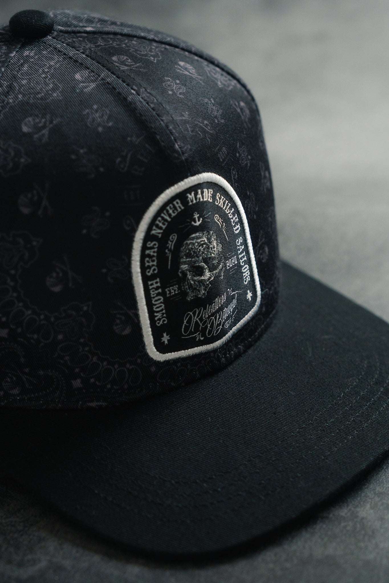 Close-up of the Skilled Sailor 5th Anniversary Snapback by Relentless Betrayal featuring a nautical-themed patch with the text Smooth Seas Never Made Skilled Sailors. The textured black cap rests on a gray surface, symbolizing resilience in overcoming challenges.