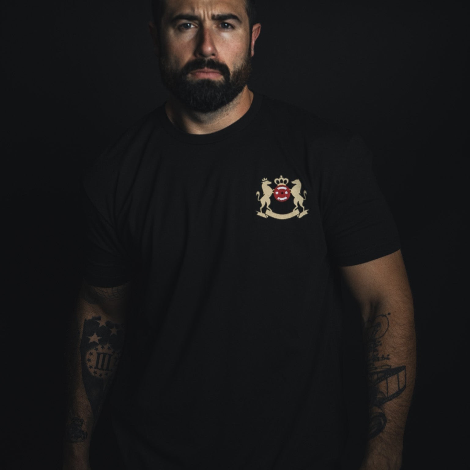 A bearded man stands against a dark backdrop, wearing the Deadlift Addiction Tee by Tango Charlie Apparel, featuring a crest with a red lion and mythical creatures. Tattoos snake down his arms as he gives you a serious look, echoing the lifter generals warning to challengers.