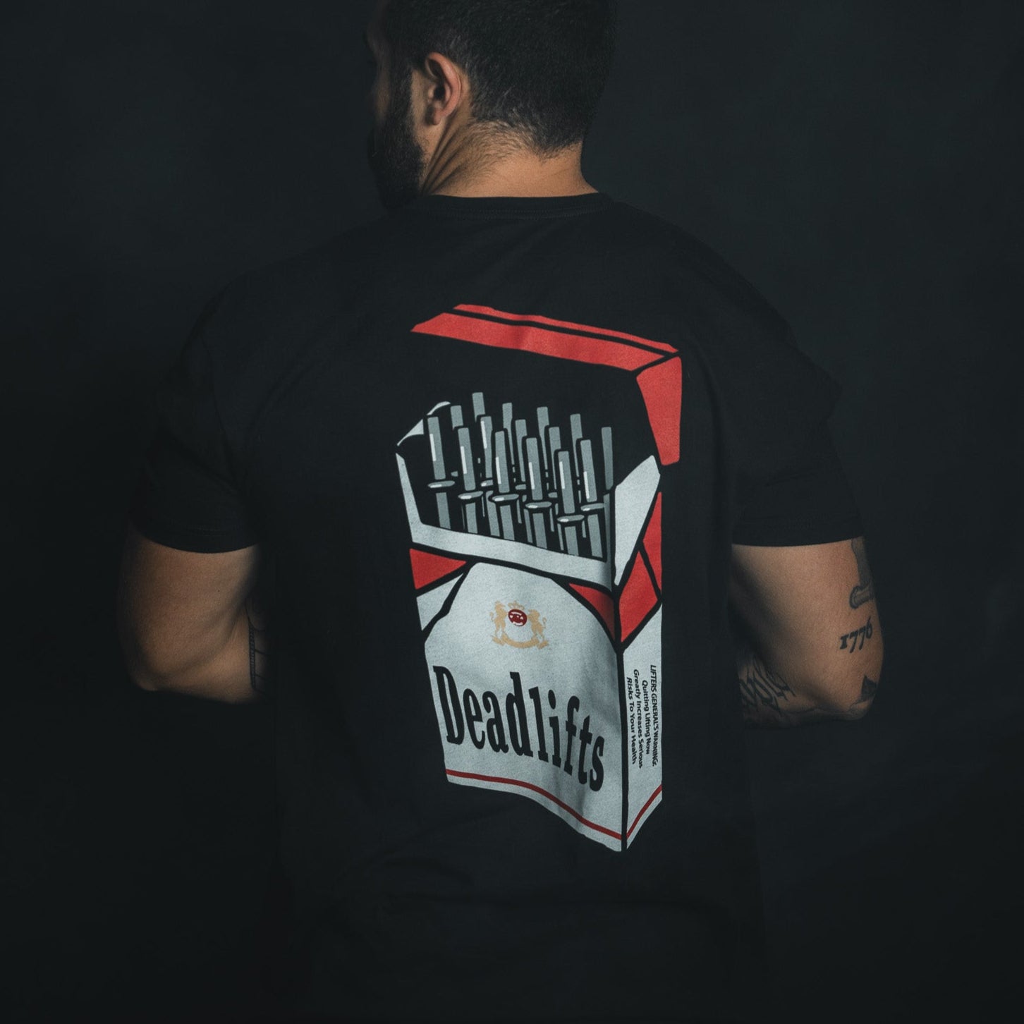 A man showcases his love for lifting in a Deadlift Addiction - Tee by Tango Charlie Apparel. The tee, with its Deadlifts cigarette pack design of mini barbells in red, white, and black, strikingly contrasts his tattooed arms against a dark background—a true lifter generals warning.