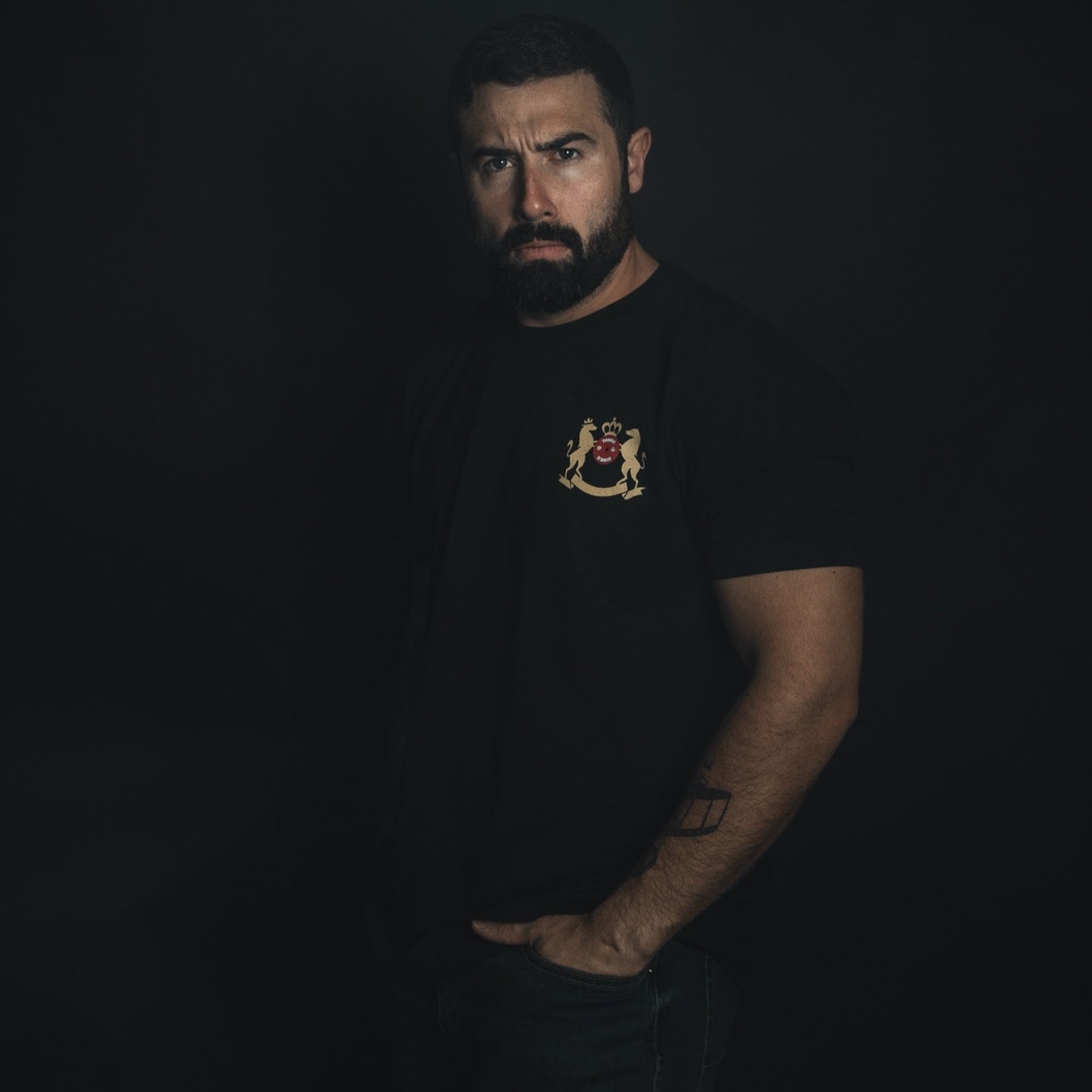 A man in a dim setting wears a Deadlift Addiction Tee by Tango Charlie Apparel featuring a chest crest. With a hand partly in his pocket and serious expression, he radiates the focused intensity of a dedicated lifter.