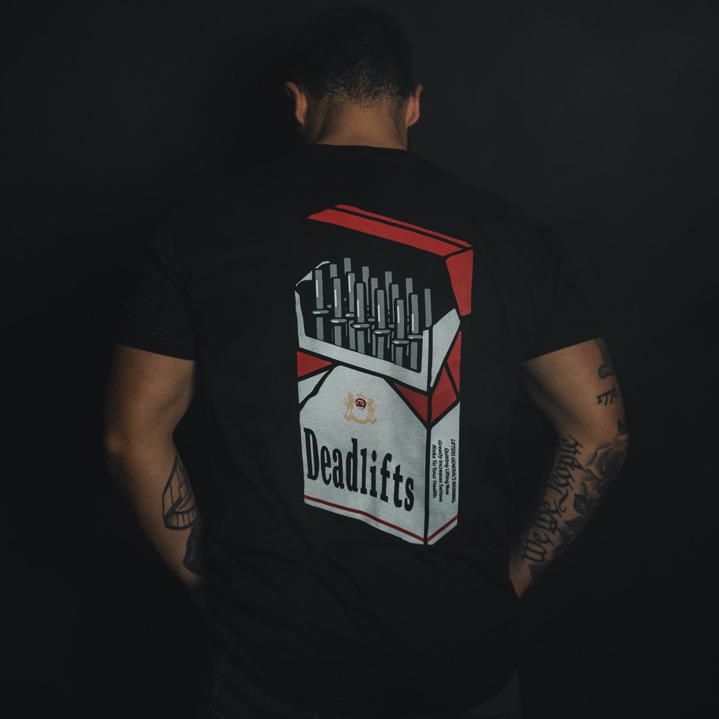 A tattooed individual turns away from the camera, wearing Tango Charlie Apparels Deadlift Addiction - Tee, a black shirt featuring a cigarette pack graphic labeled Deadlifts with a humorous lifter generals warning.