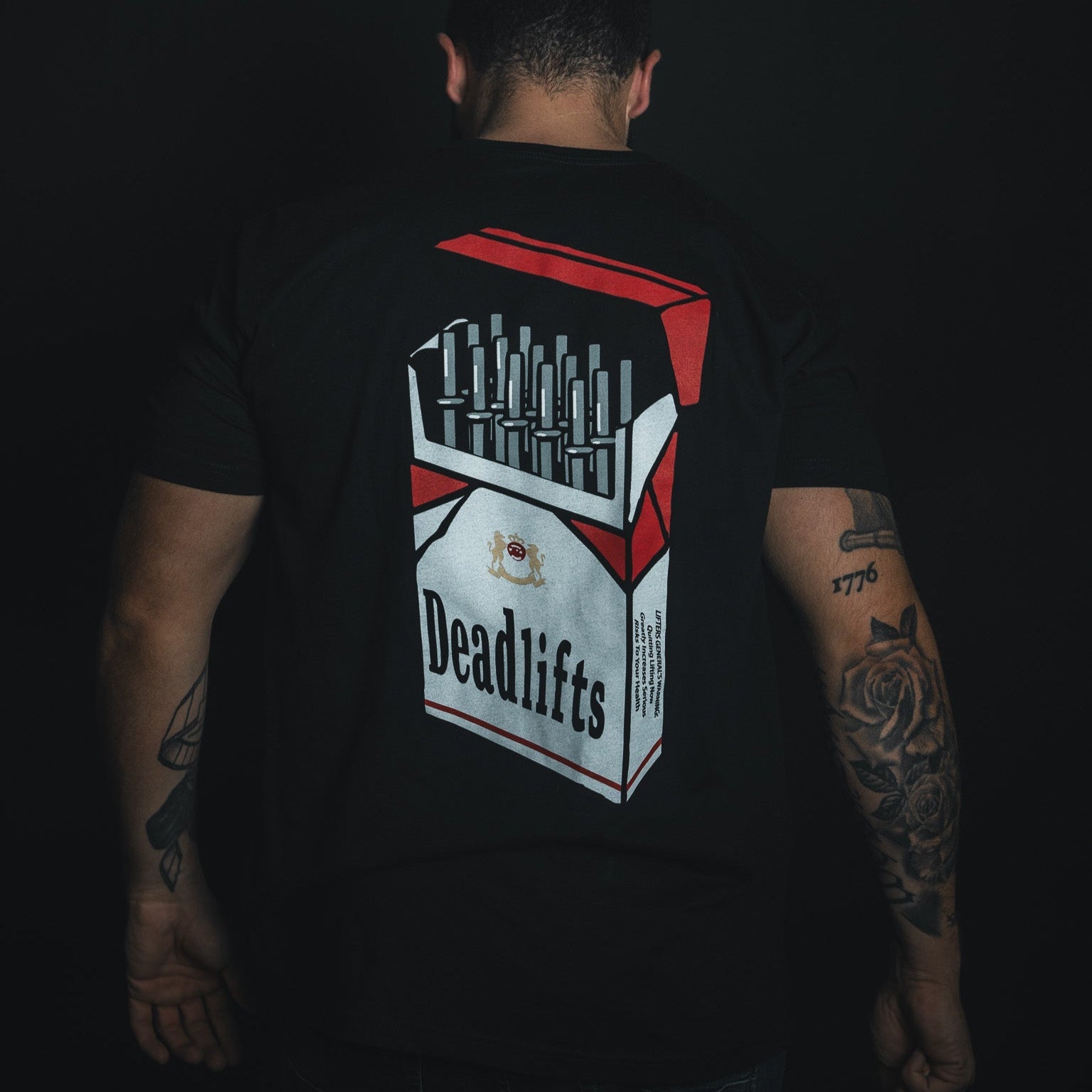 A person faces away wearing the Deadlift Addiction Tee from Tango Charlie Apparel, showcasing a cigarette pack graphic labeled Deadlifts, filled with barbells. Tattoos on their arms warn of the deadlift lifestyles power and allure.