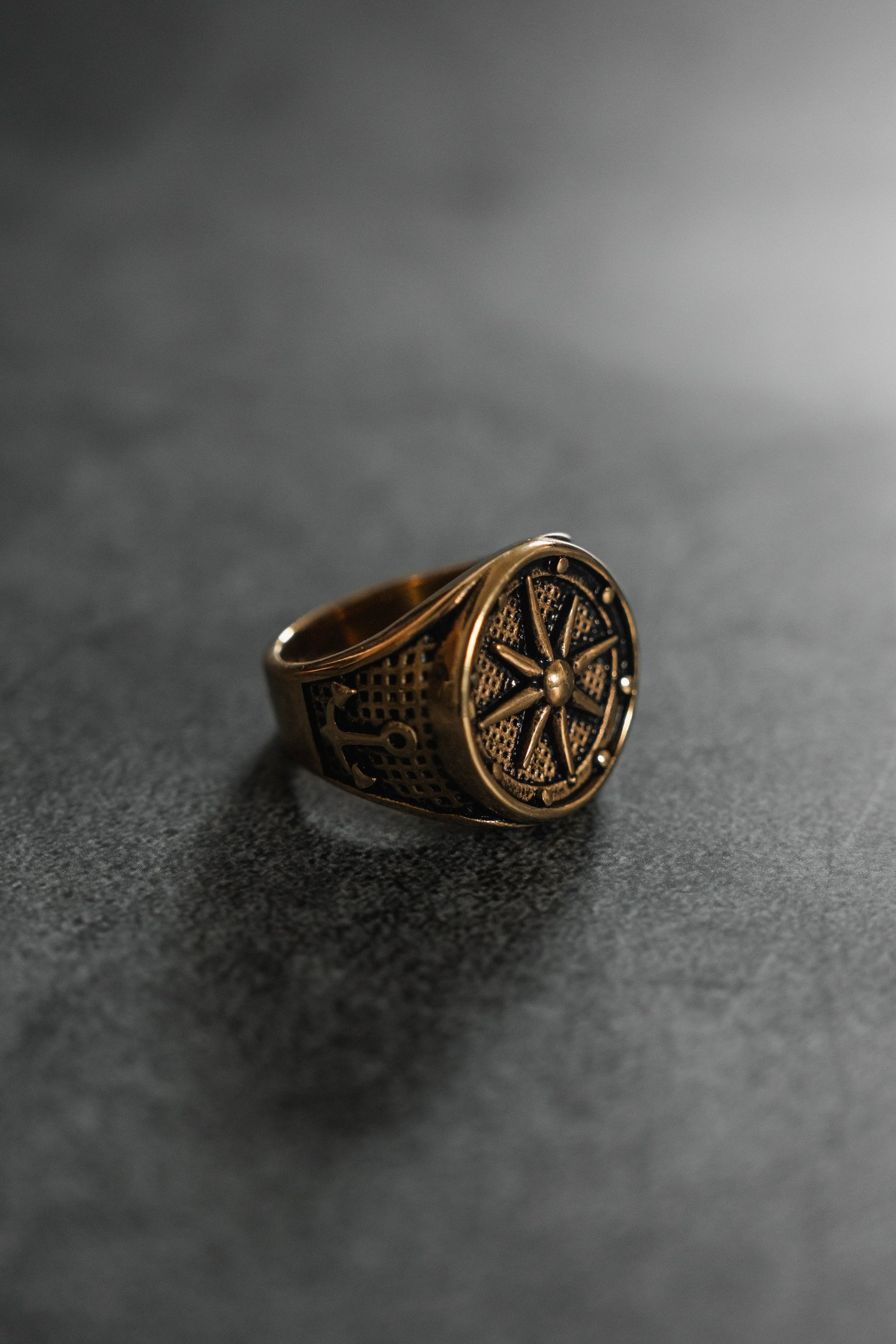 A Neptunes Compass Ring from Relentless Betrayal, featuring an ornate, bronze design with a compass-like emblem, rests on a dark textured surface. The lighting highlights its intricate details and casts subtle celestial-inspired shadows.