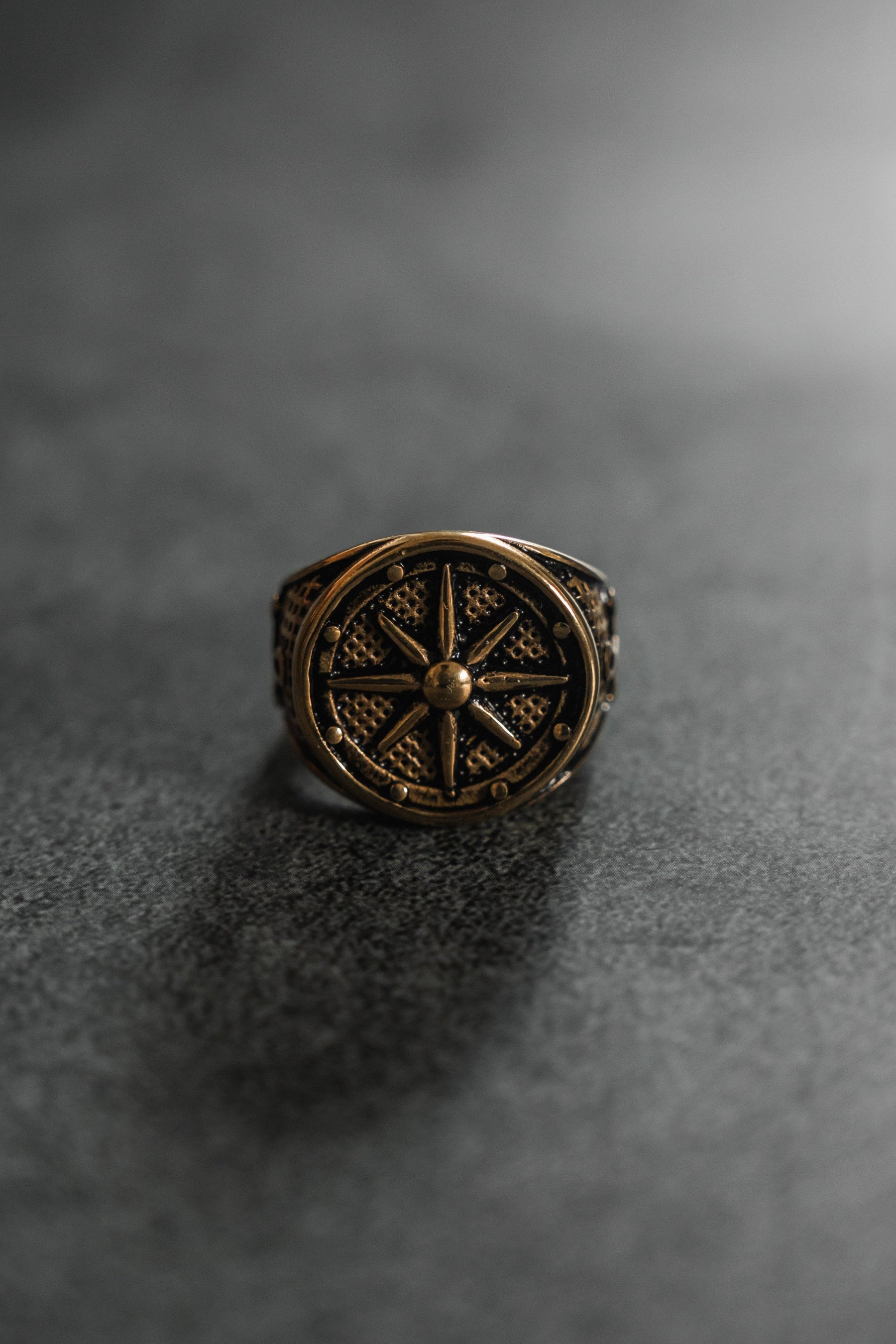 Relentless Betrayal presents Neptunes Compass Ring, a bronze masterpiece with intricate engravings and a compass-like design on a dark, textured surface. Celestial inspiration shines through as it stands upright, casting a subtle shadow.