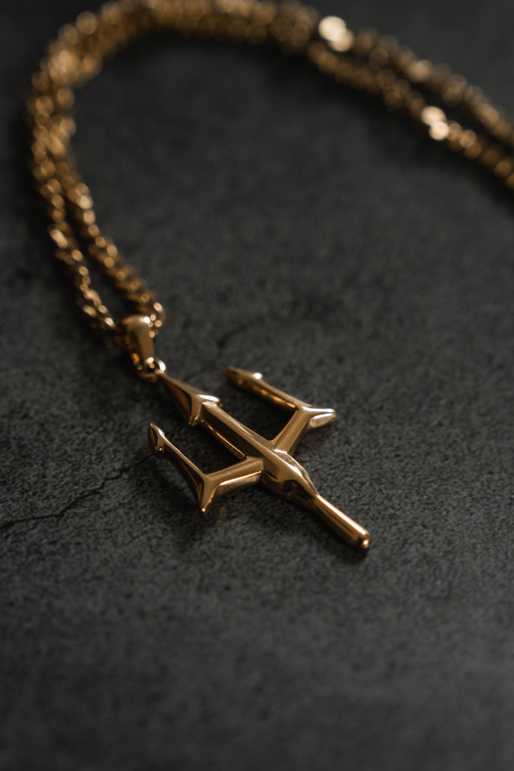 Against a dark, textured backdrop, Relentless Betrayals Poseidons Trident Necklace with its gold, trident-shaped pendant on a matching chain exudes maritime strength and embodies the polished elegance of Greek mythology jewelry.