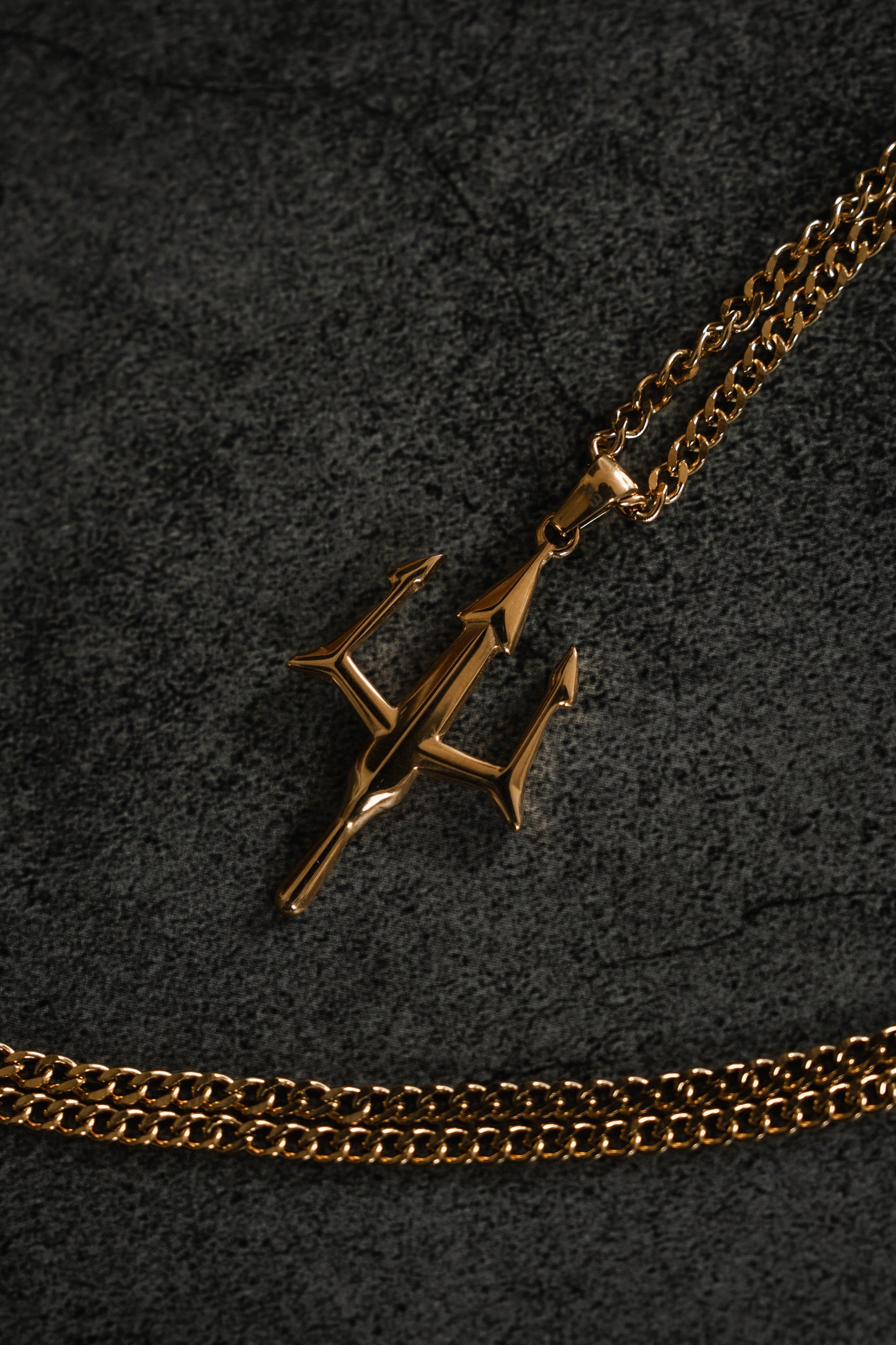 A gold pendant from Relentless Betrayals Poseidons Trident Necklace shines on a dark, textured surface, with another chain partially visible beneath, embodying the maritime strength of Greek mythology jewelry.