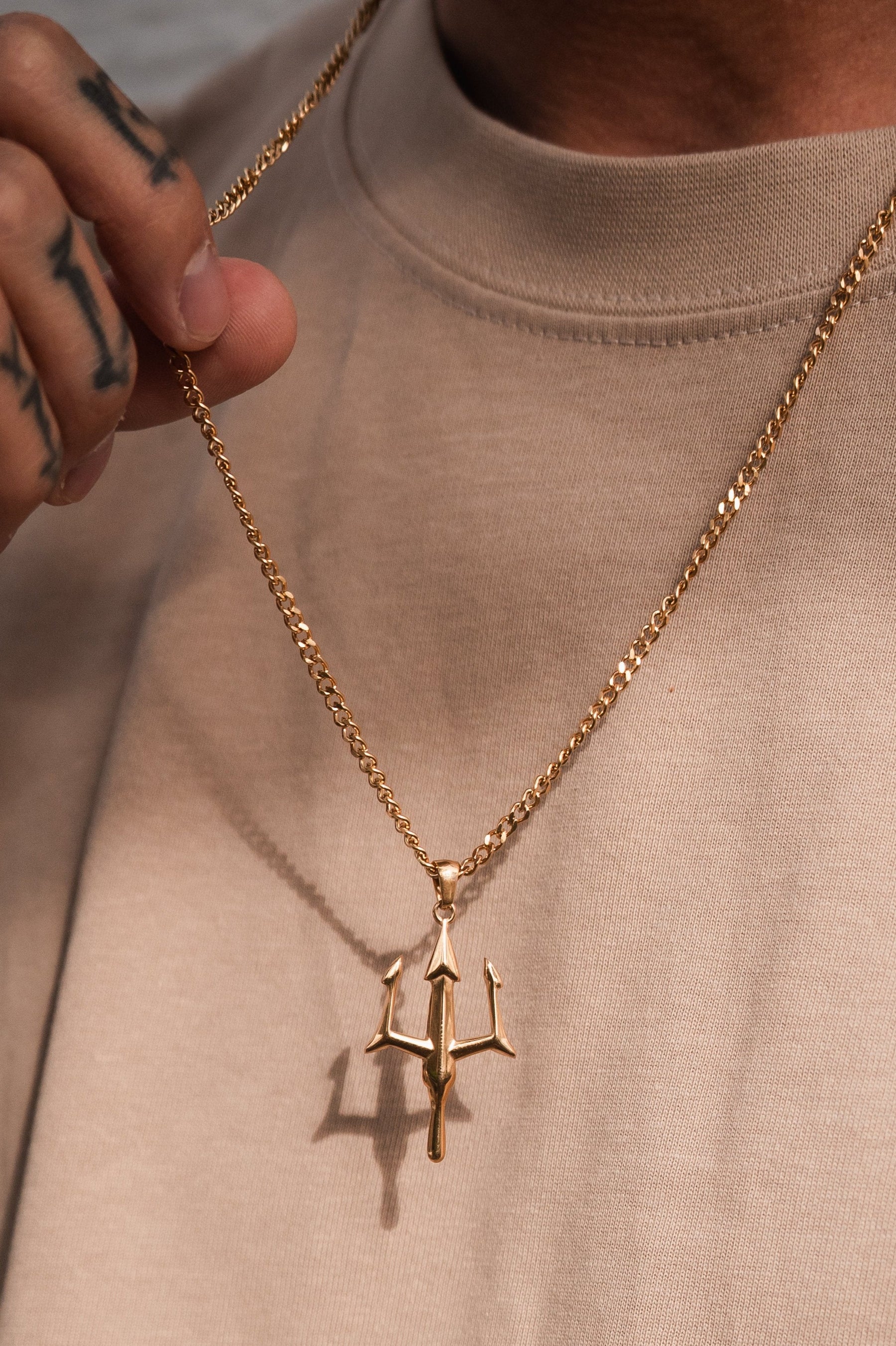 A person in a beige shirt holds the Poseidons Trident Necklace by Relentless Betrayal, their tattooed fingers gently clutching the chain. This Greek mythology jewelry gleams with maritime strength, the trident-shaped pendant showcasing its captivating metallic allure.