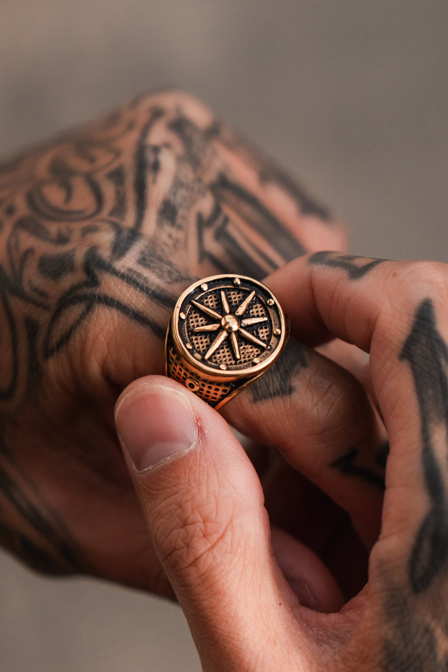 A tattooed hand elegantly holds the Neptunes Compass Ring by Relentless Betrayal, a gold piece with a celestial design. Its intricate lines resemble deep blue sapphires against an out-of-focus background.