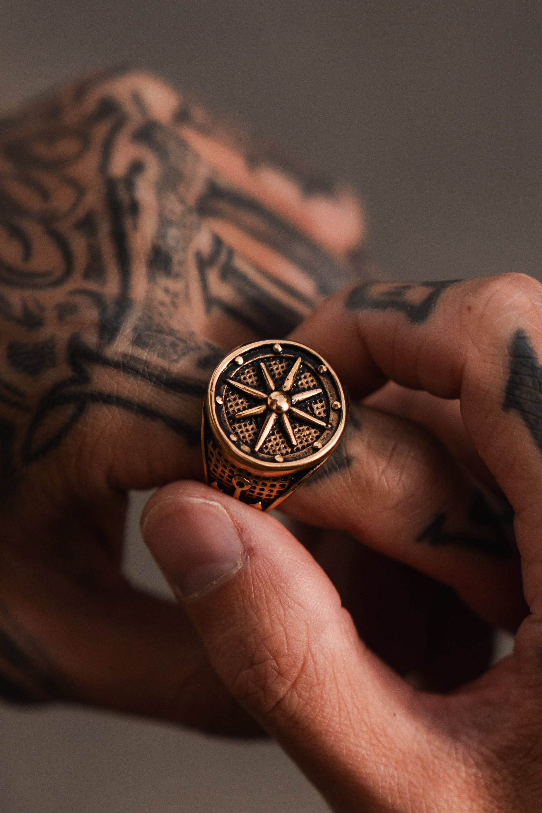 A tattooed hand holds the Neptunes Compass Ring by Relentless Betrayal, showcasing a gold piece with a cross design and deep blue sapphires, set against a softly blurred background.