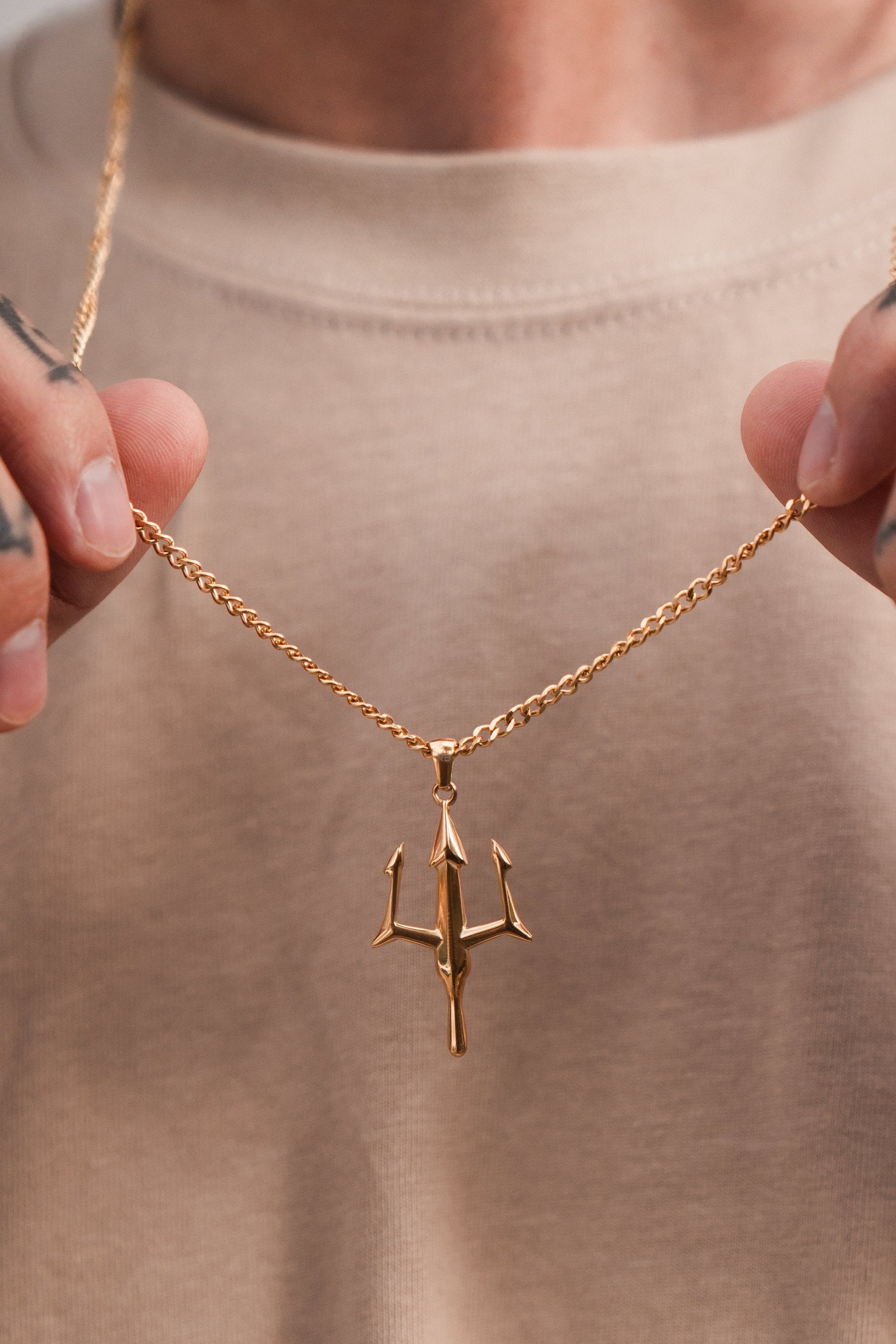 Close-up of Poseidons Trident Necklace by Relentless Betrayal, symbolizing maritime strength. The gold piece shines against a beige shirt amid finger tattoos, with a blurred background evoking Greek mythology jewelrys mystique.
