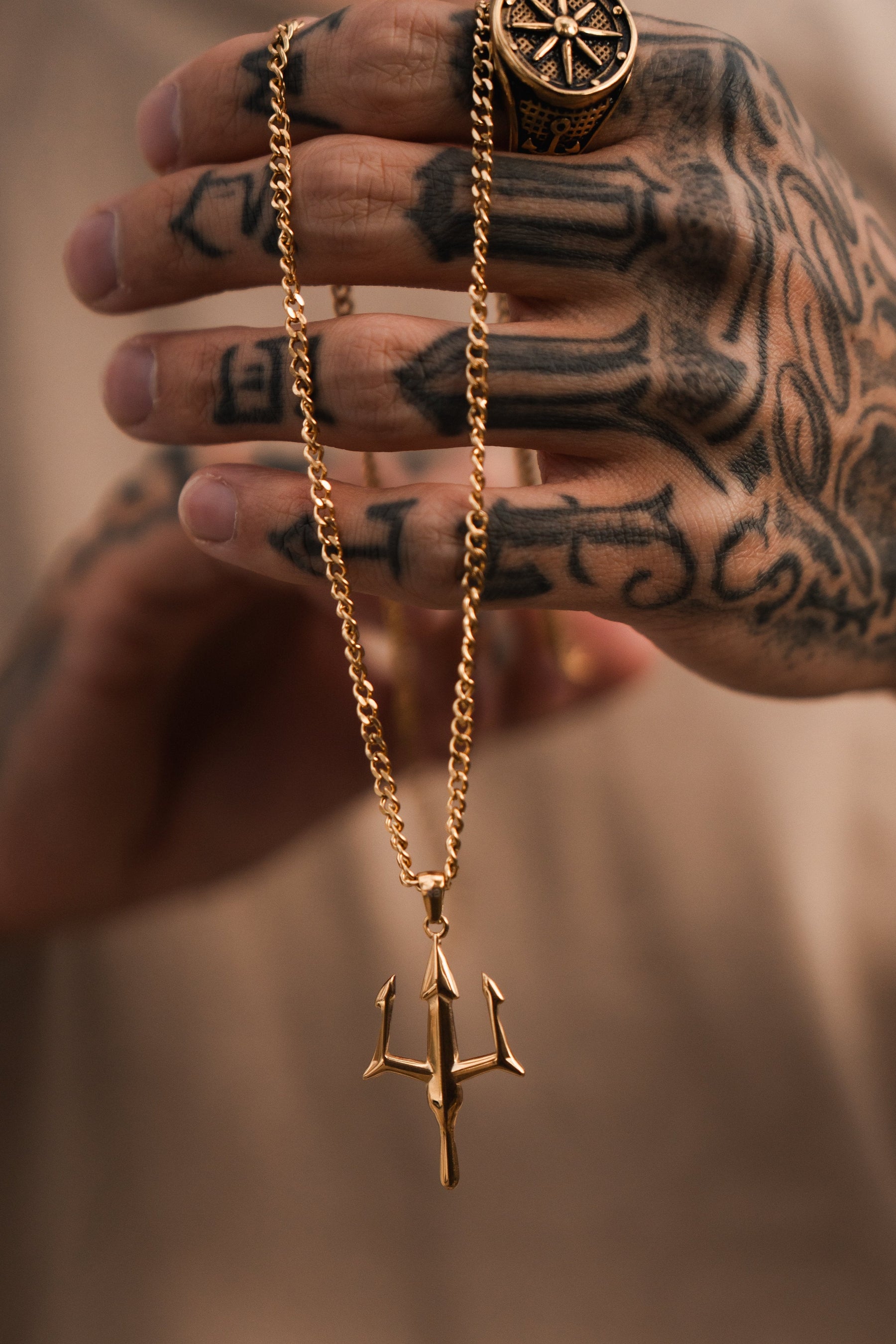 A tattooed hand holds Relentless Betrayals Poseidons Trident Necklace, symbolizing maritime strength. The person wears a large ornate ring, while the softly blurred background accentuates the intricate Greek mythology jewelry and striking tattoos.