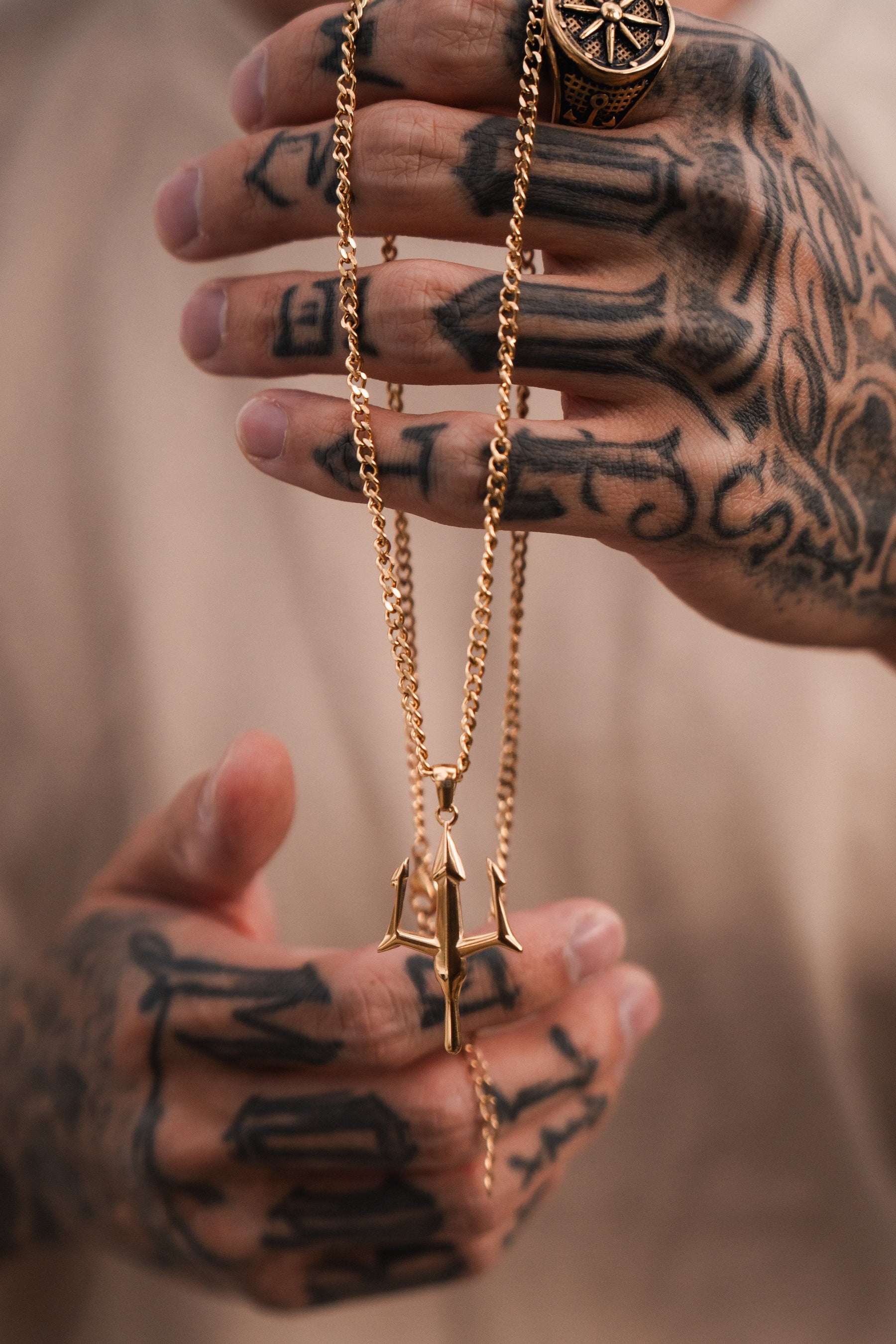 Tattooed hands clasp a Poseidons Trident Necklace by Relentless Betrayal, highlighting the maritime strength of Greek mythology jewelry. A ring with an intricate pattern adorns a finger against a softly blurred background, adding mystery.