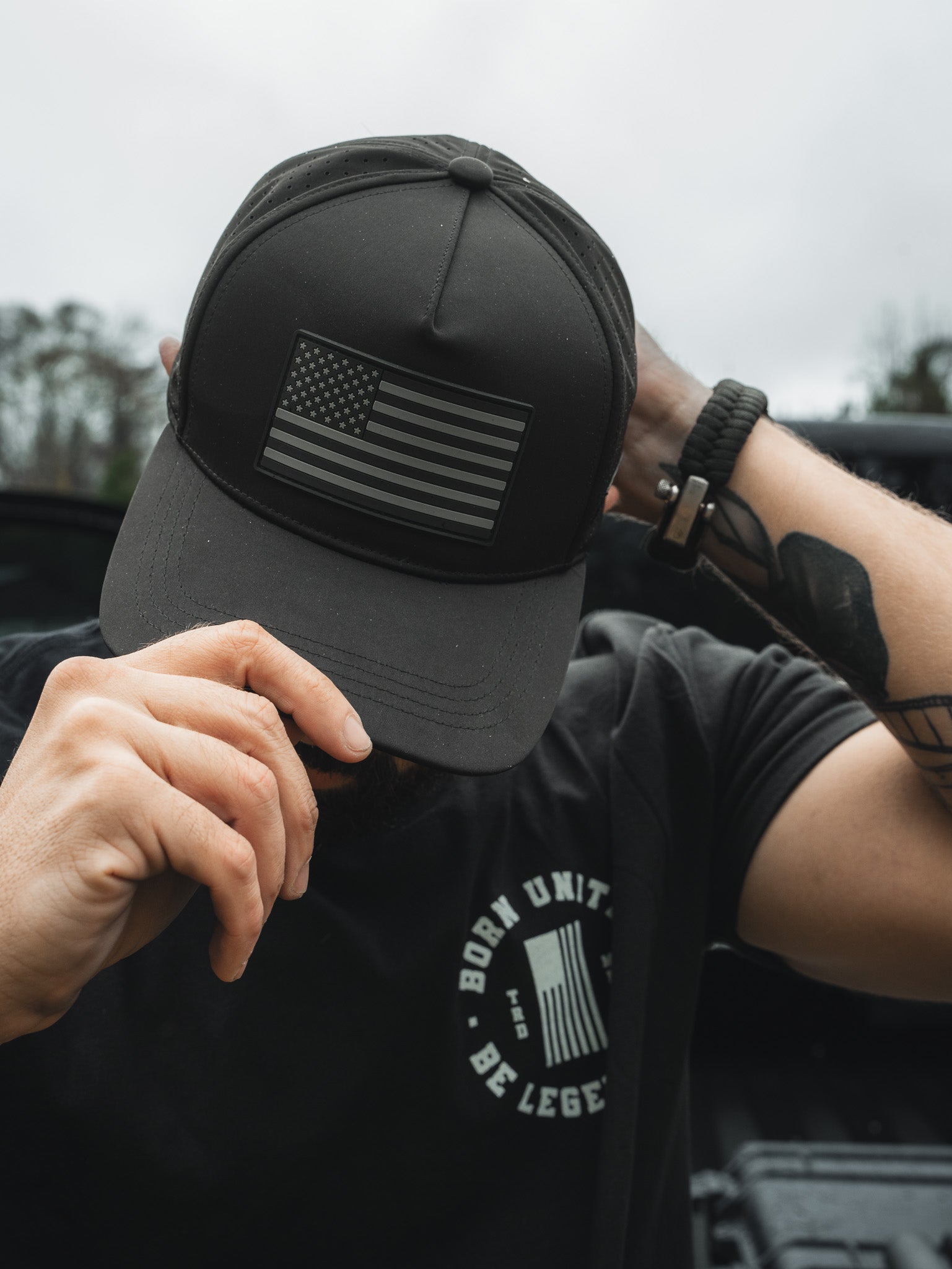 Person adjusts a Born United Premium Flag Snapback - Blackout, displaying a monochrome American flag patch. They sport a black T-shirt, watch, and visible tattoos on their arm. The blurred backdrop features trees.