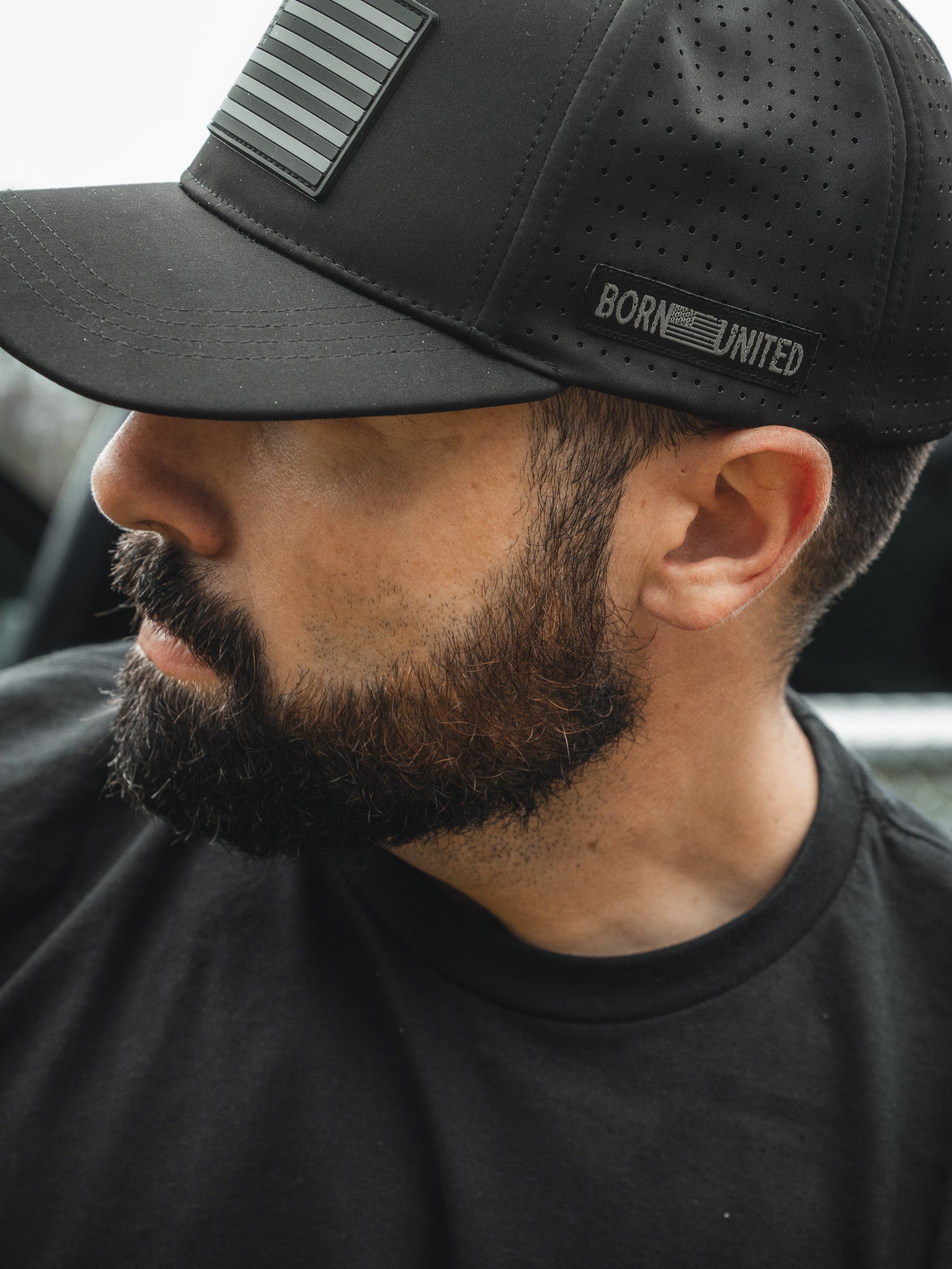 A bearded man wears a Born United Premium Flag Snapback - Blackout by Born United Private Label and a T-shirt, featuring perforations, BORN UNITED, and a stylized American flag near the brim, with his face partially turned from the camera.