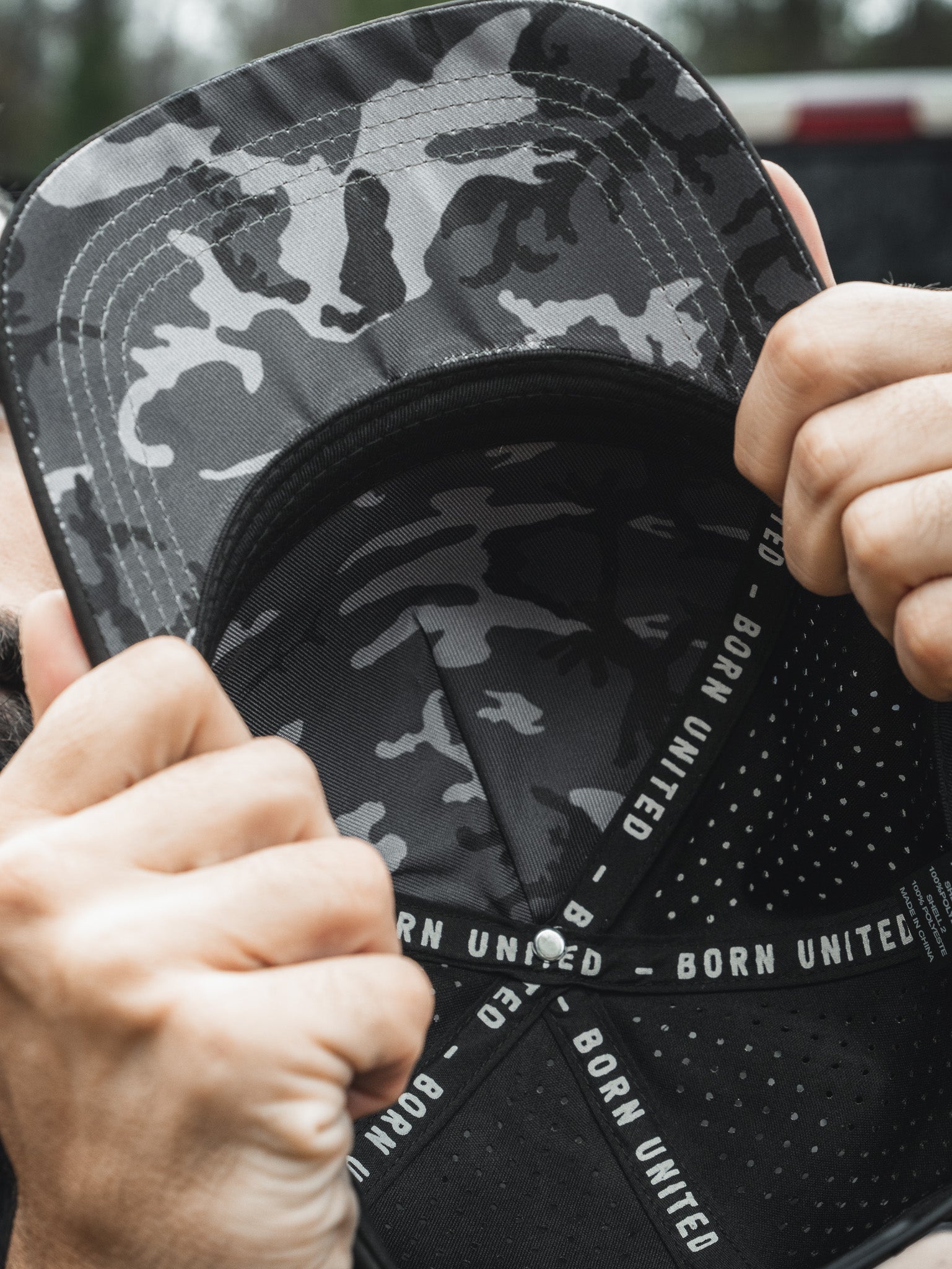 A person grips the brim of a camo-patterned Born United Premium Flag Snapback - Blackout, subtly concealing their face. The inner band reads BORN UNITED, while the blurred background accentuates the caps intricate details.