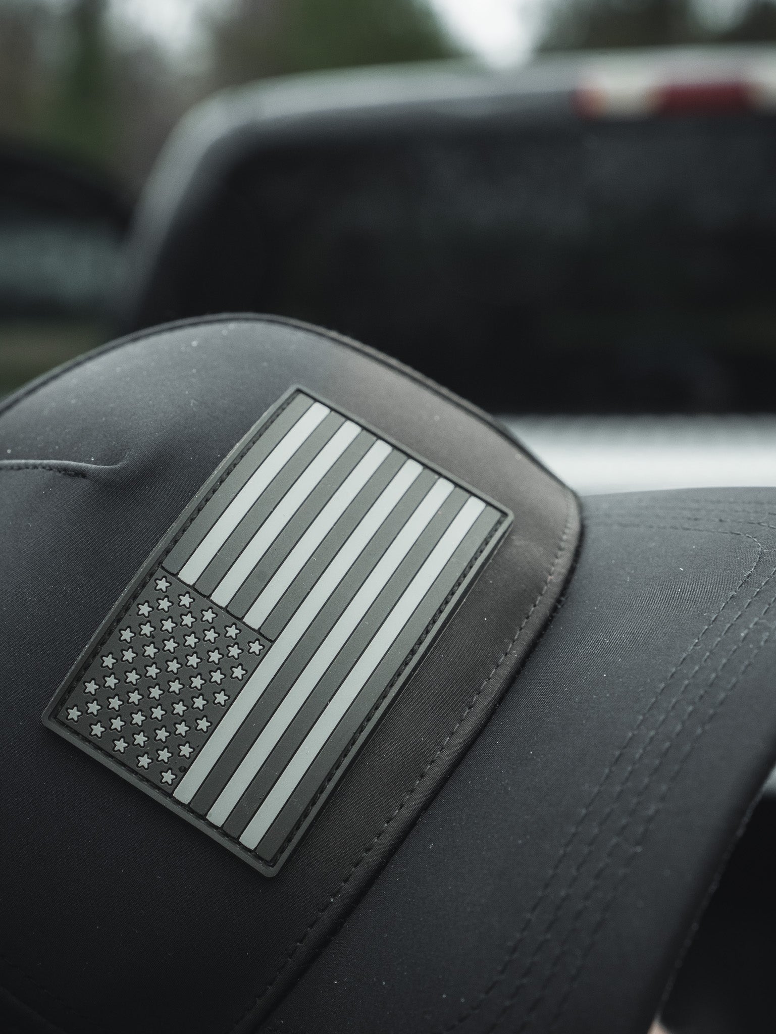 Close-up of the Born United Premium Flag Snapback - Blackout by Born United Private Label, showcasing a black-and-white American flag patch on grey. The caps brim is in focus with a blurred vehicle in the background.