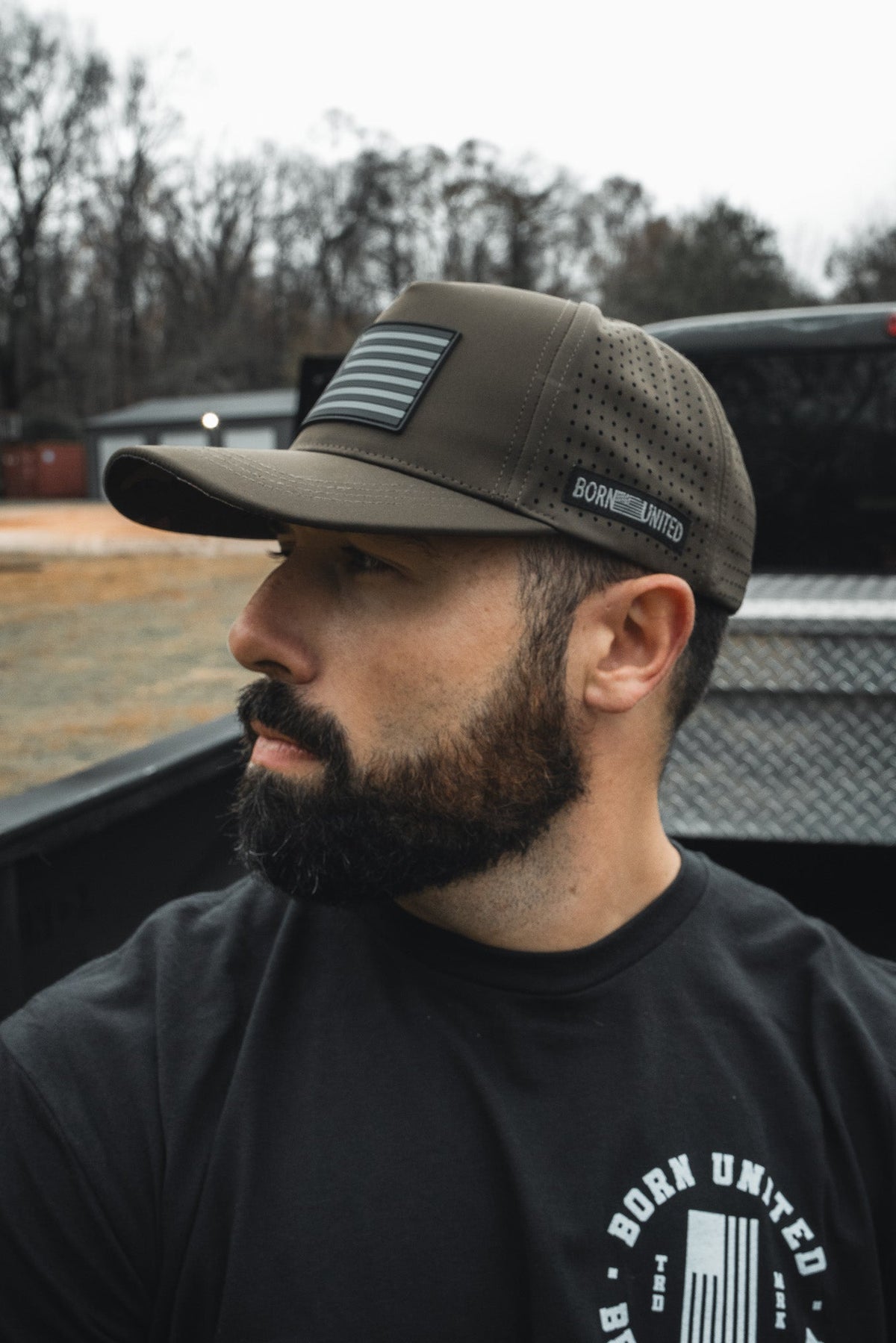 A bearded man in a black T-shirt and an olive green "Legend - Saved" baseball cap by Born United Private Label with an American flag patch sits in a pickup truck bed, looking to the side. Trees and a building blur in the background.