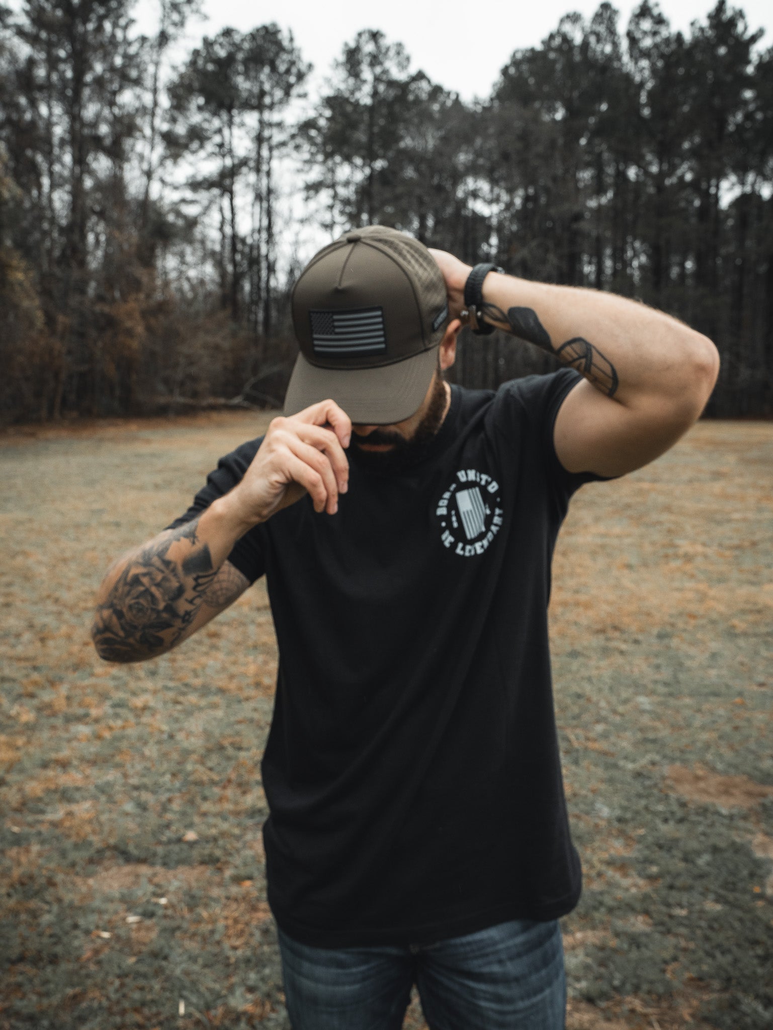 A man in a black t-shirt and camo cap stands in a grassy field, trees behind him. Tattoos cover his right arm as he adjusts the Born United Premium Flag Snapback - Olive featuring a black and white American flag design, with his left hand.