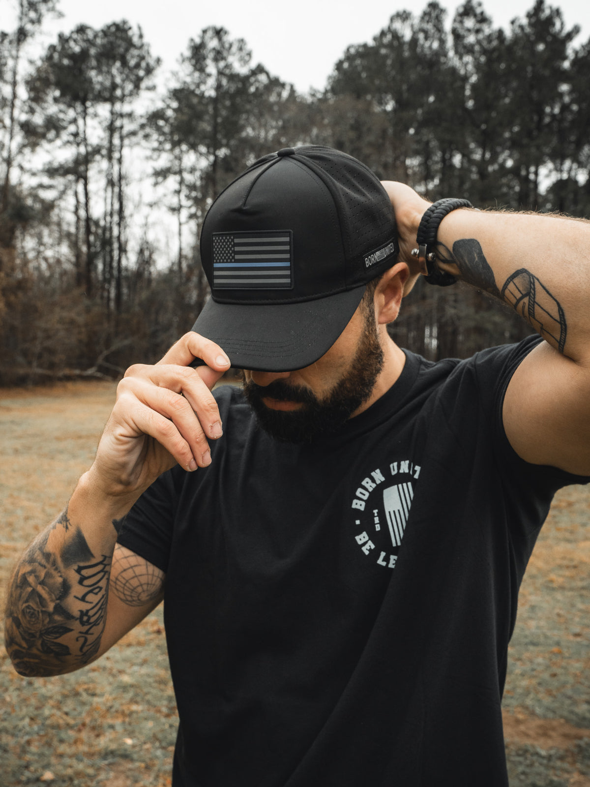 A man outdoors adjusts his Born United Premium Flag Snapback - TBL, a black Born United Private Label cap with an American flag, while wearing a black T-shirt with text. His arm tattoos are visible against tall trees in the background.