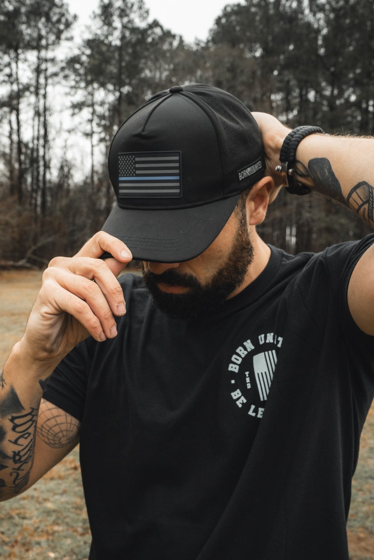 A man outdoors adjusts his Born United Premium Flag Snapback - TBL, a black Born United Private Label cap with an American flag, while wearing a black T-shirt with text. His arm tattoos are visible against tall trees in the background.